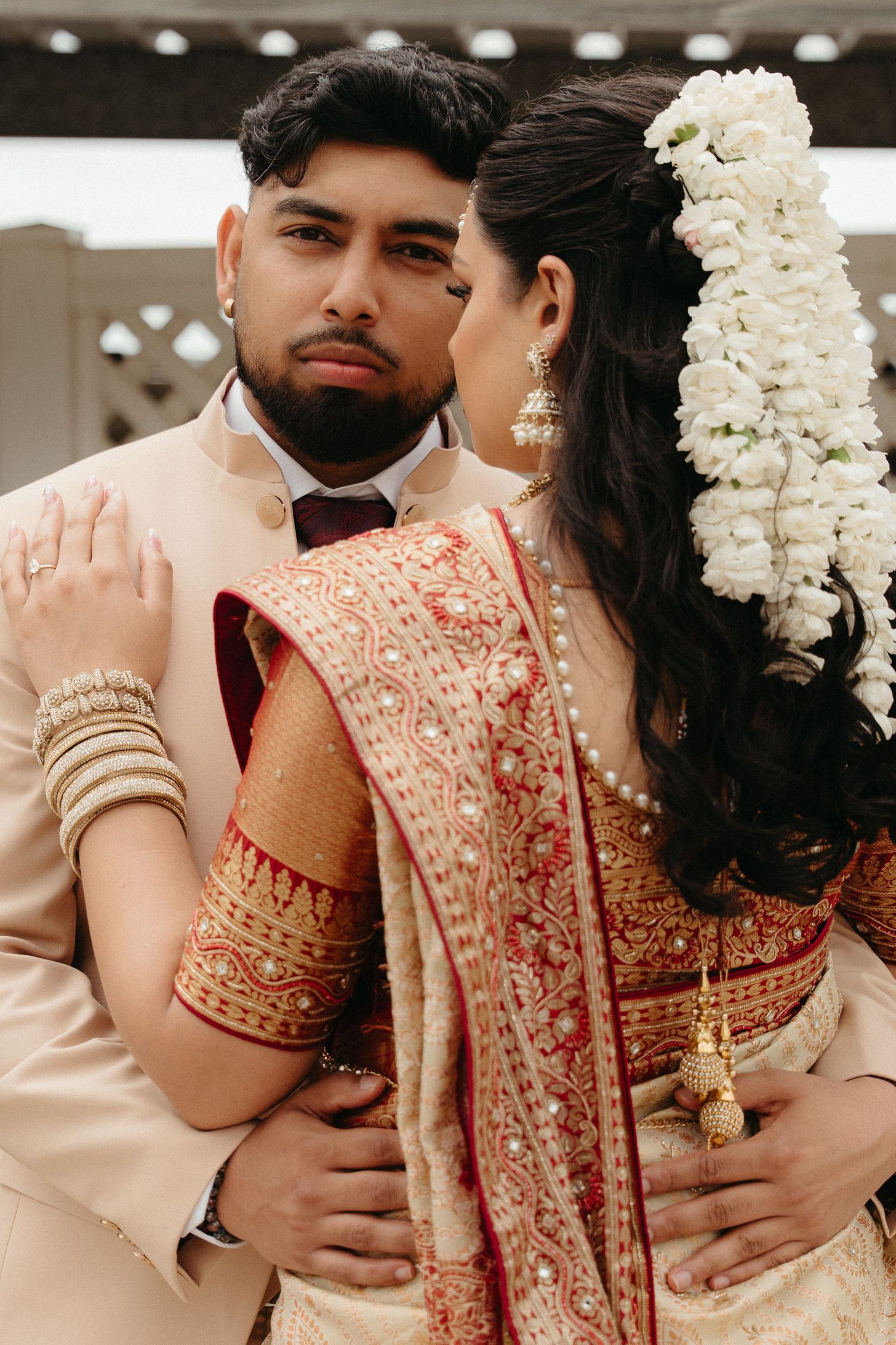 Close-up of couple in traditional South Asian wedding attire with beige suit and red embroidered sari with white floral hair piece.