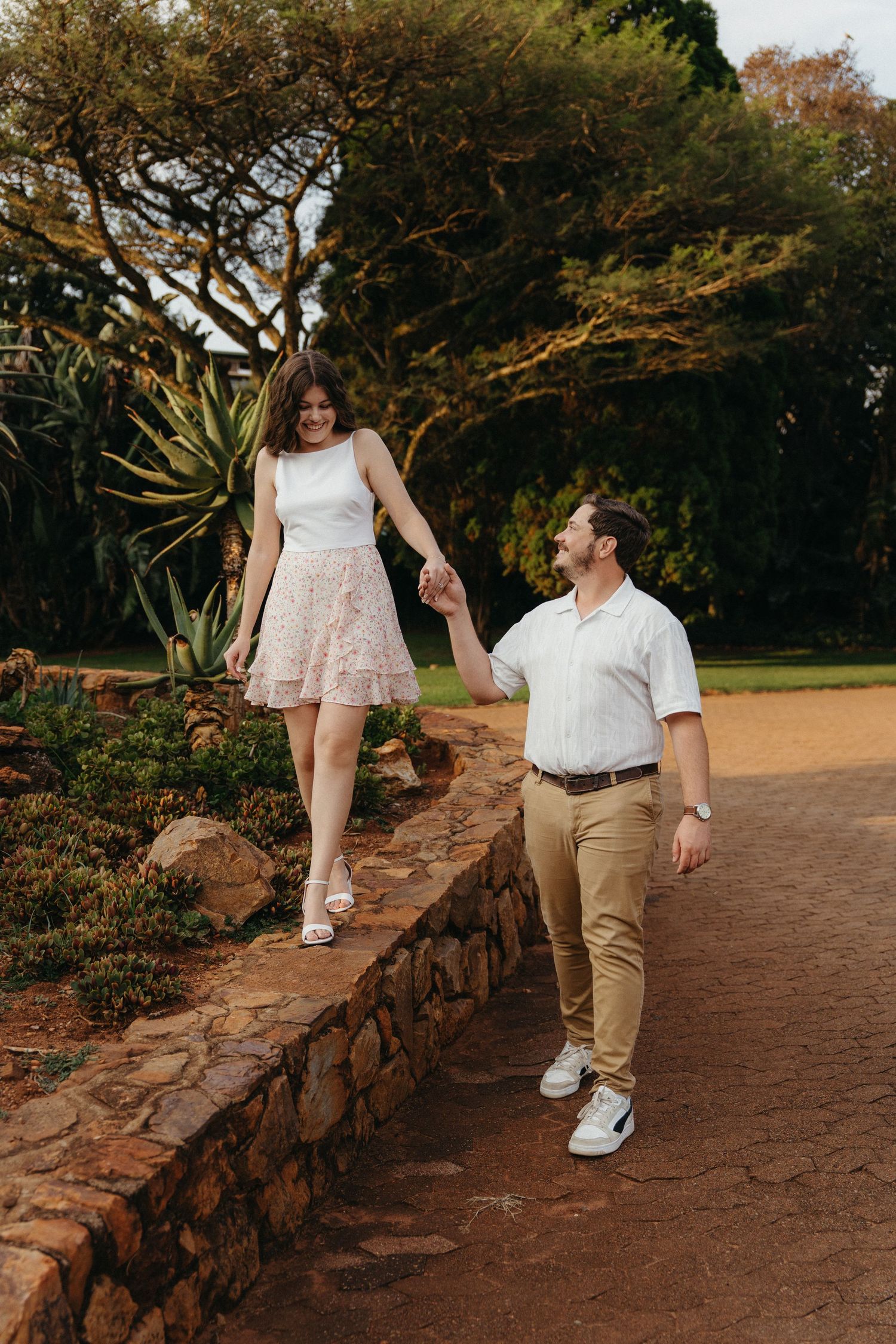 Couple holding hands walking along a stone garden path surrounded by lush greenery and trees.