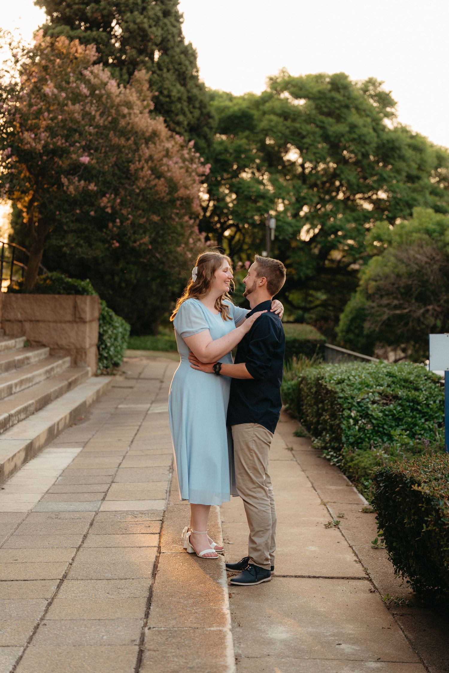 A couple in light blue and khaki share a romantic moment on stone steps surrounded by greenery at sunset.