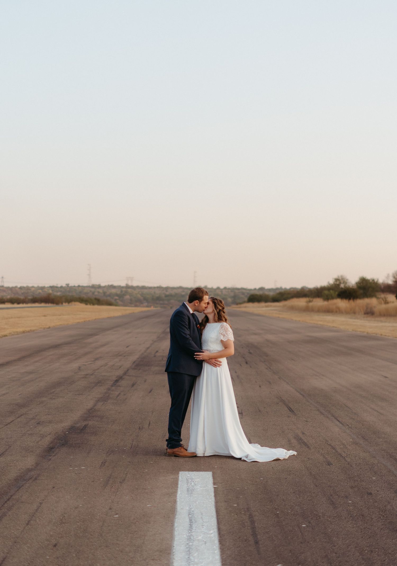 A romantic moment shared on an empty runway with planes flying overhead at dusk.