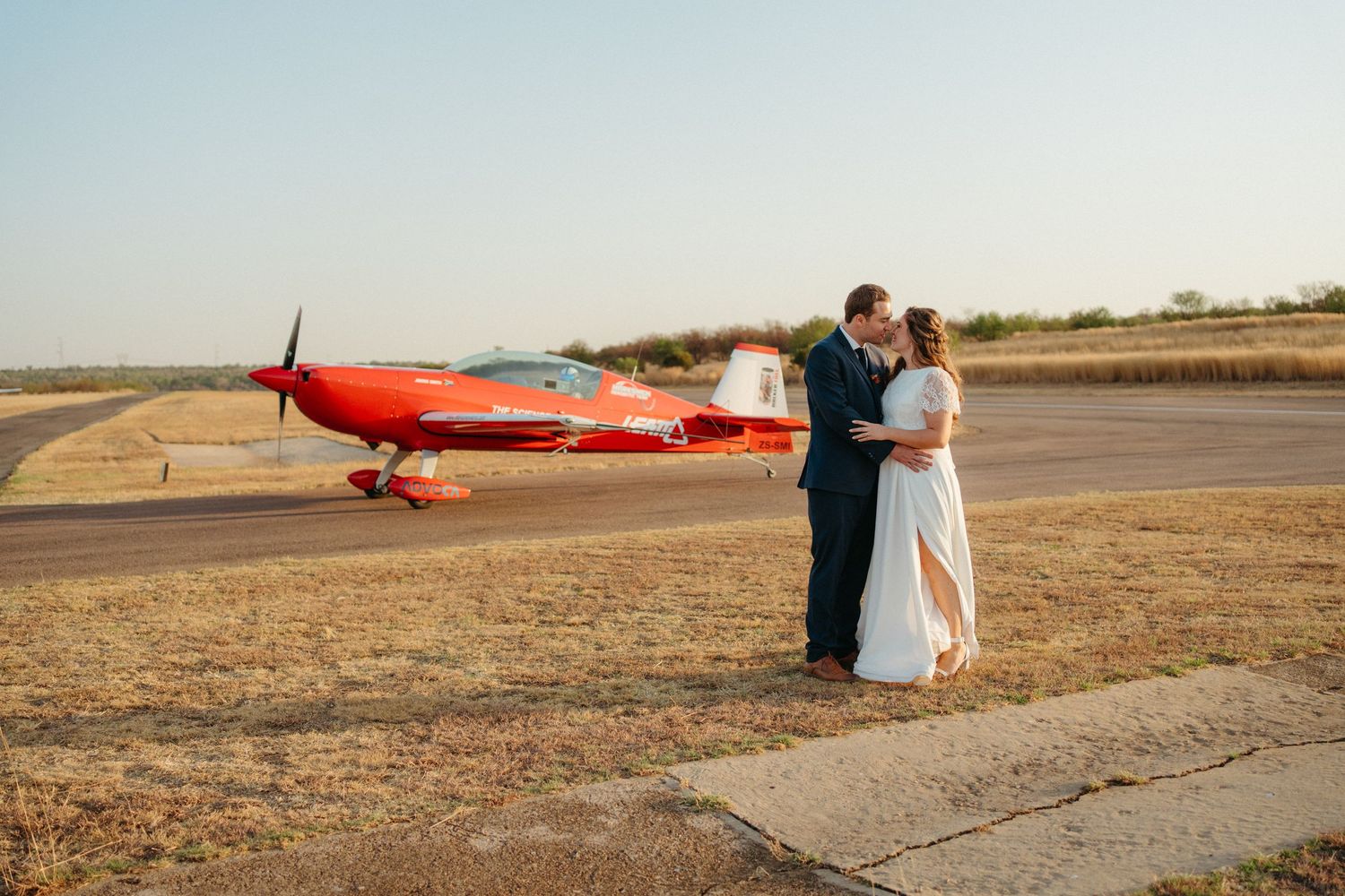 A couple embraces near a bright red vintage airplane on a dusty airfield.
