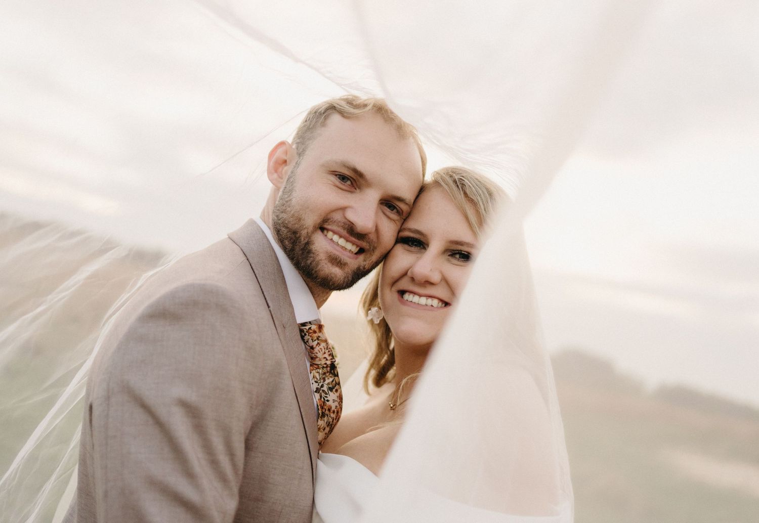 Wedding couple smiling together under flowing veil at outdoor ceremony against cloudy sky.