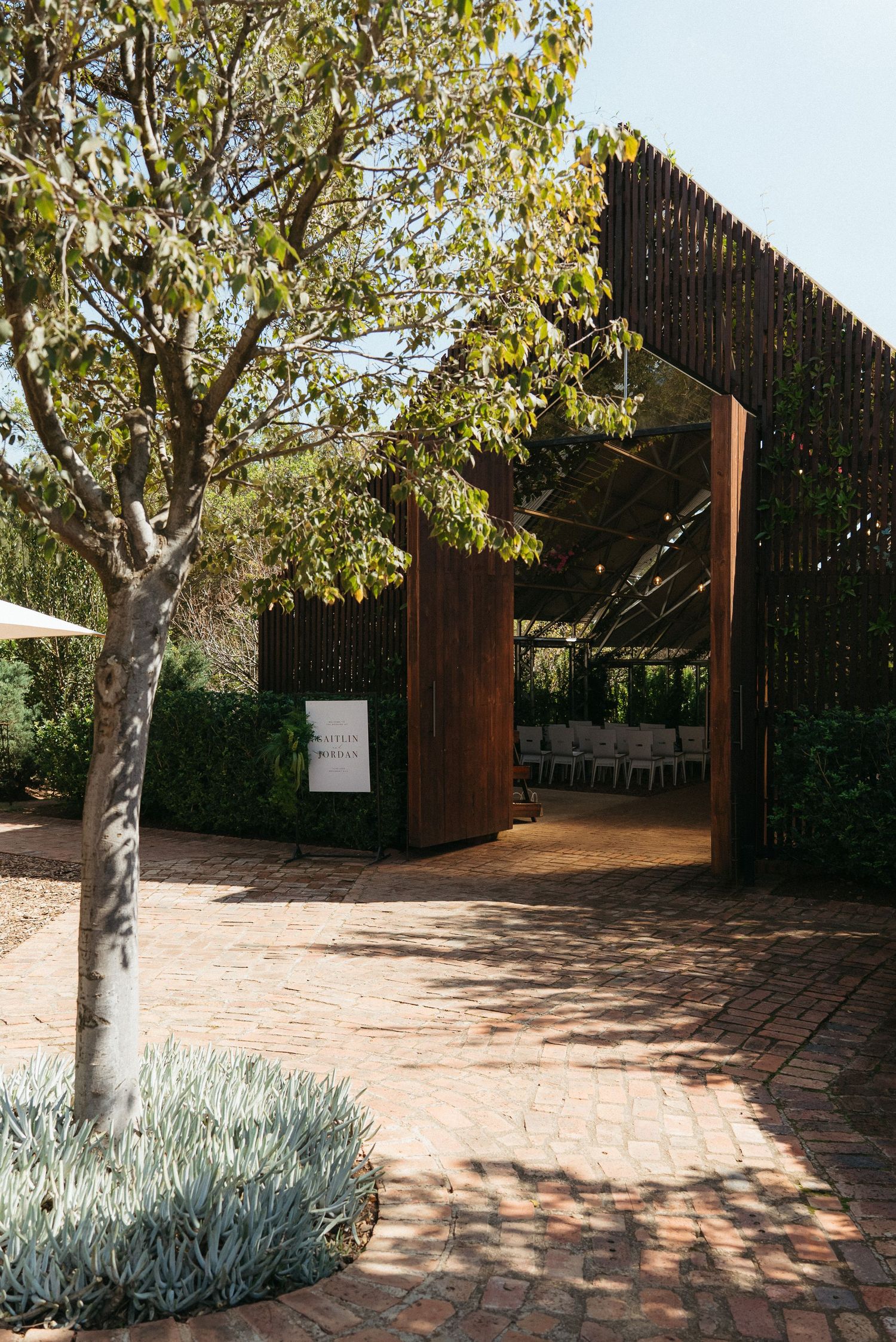 Rustic outdoor wedding venue with wooden slat walls, brick pathway, and white chairs visible through open entrance.