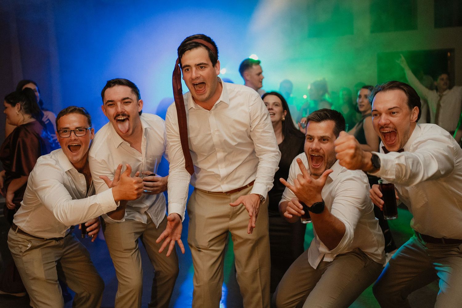 Groomsmen celebrating on the dance floor at a wedding reception with colorful lights.