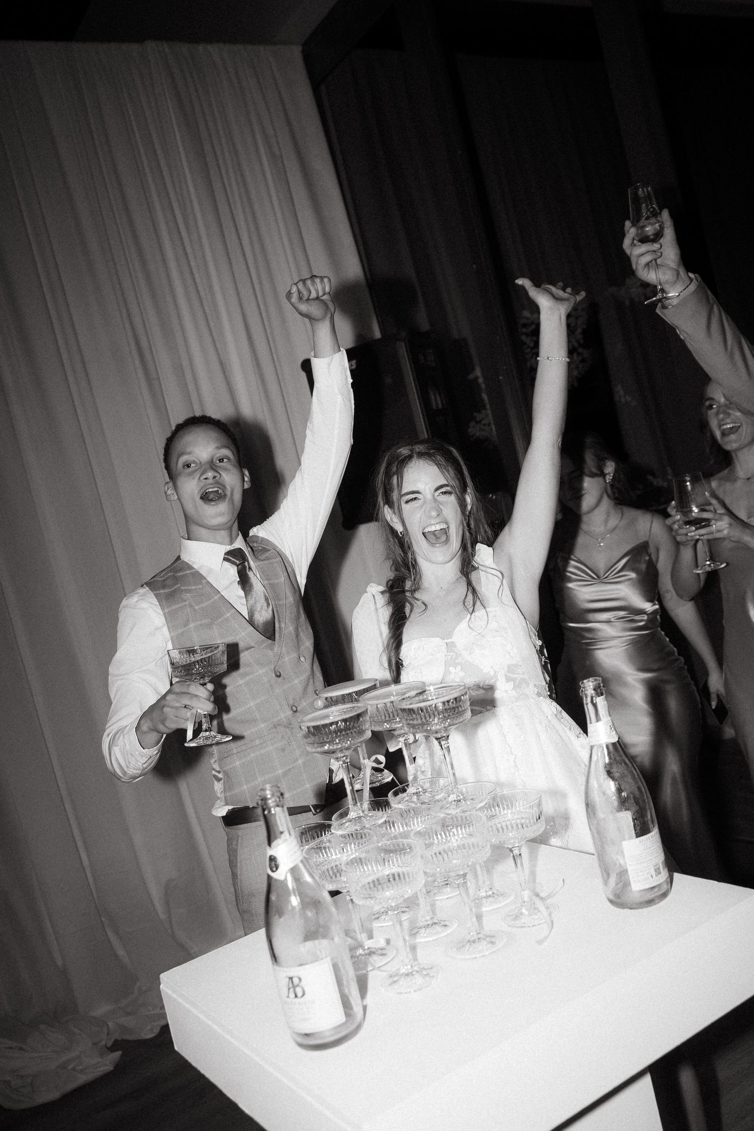 Joyful bride and groom celebrating at wedding reception with champagne tower, arms raised in excitement, black and white photo.