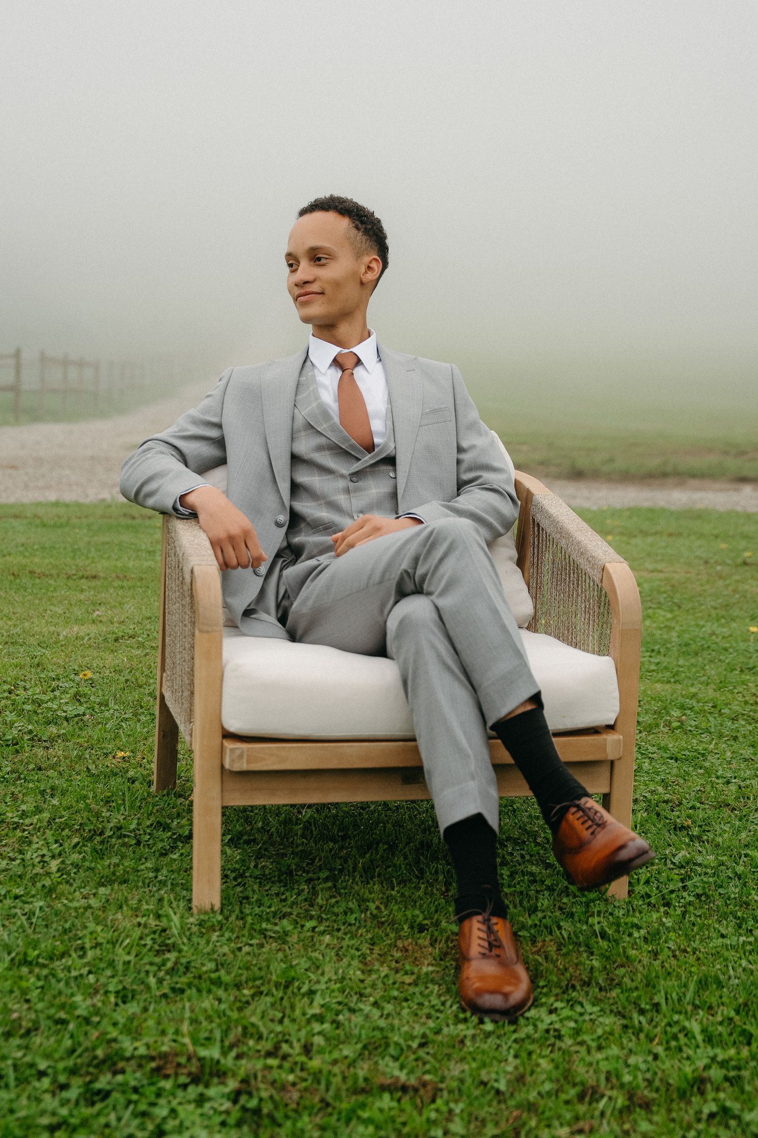 A groom in a gray suit and brown shoes sits relaxed in a wooden chair on a misty green lawn at Tafika Polo Farm.