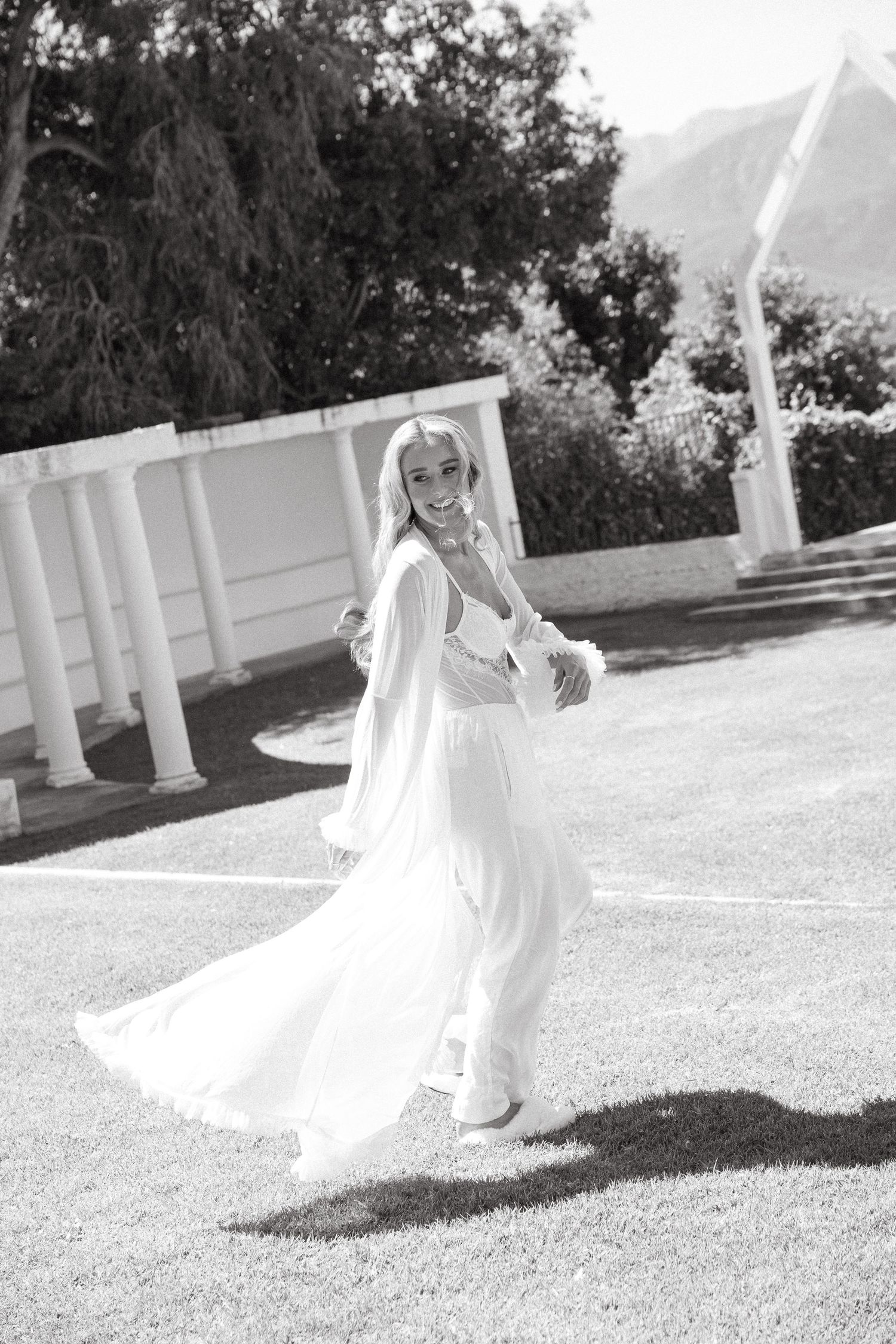 Smiling bride in a flowing white gown twirls on a sunlit lawn near white columns, with trees and mountains in the background.