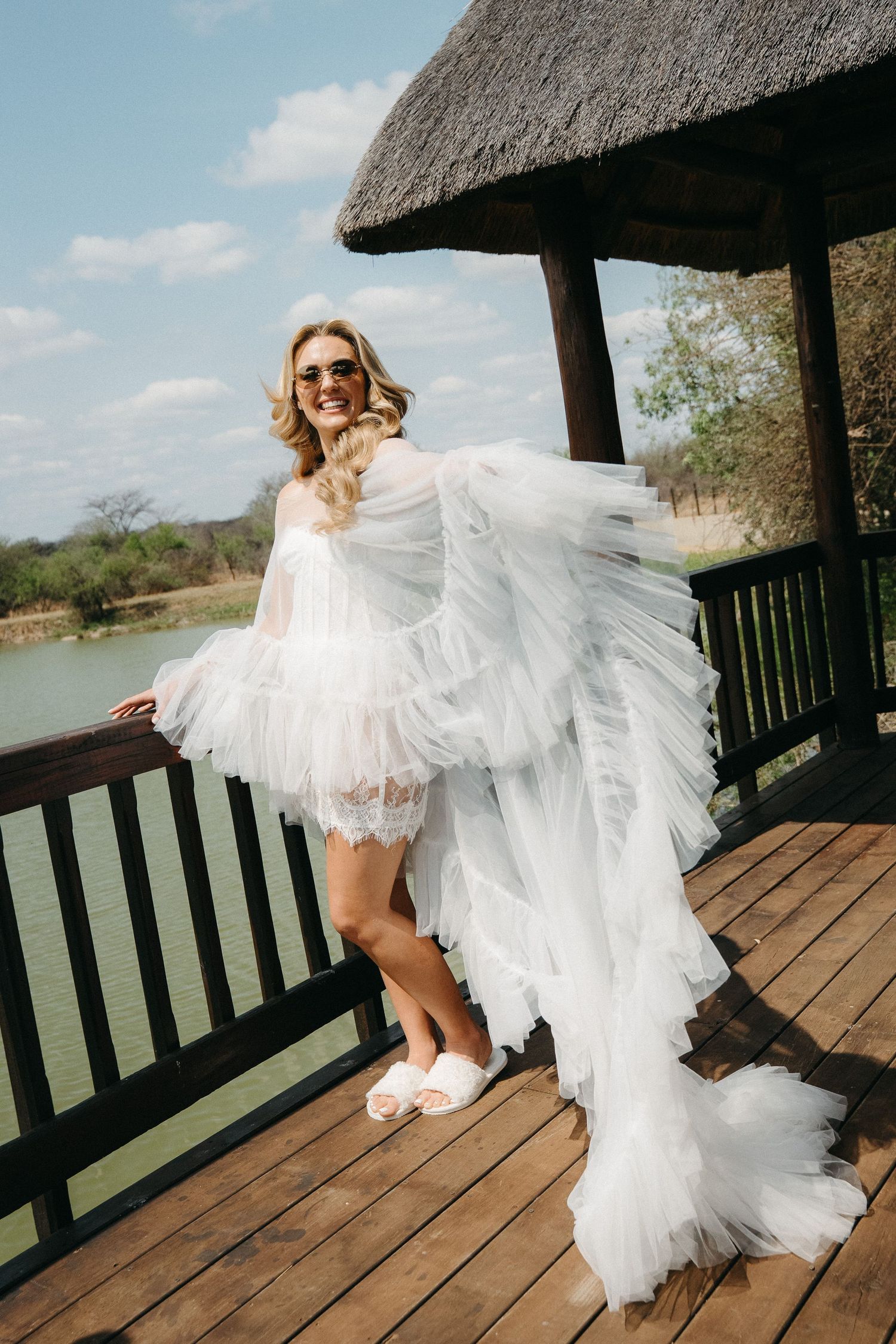 Bride in white tulle robe and sunglasses posing on wooden deck overlooking a lake at a safari lodge.