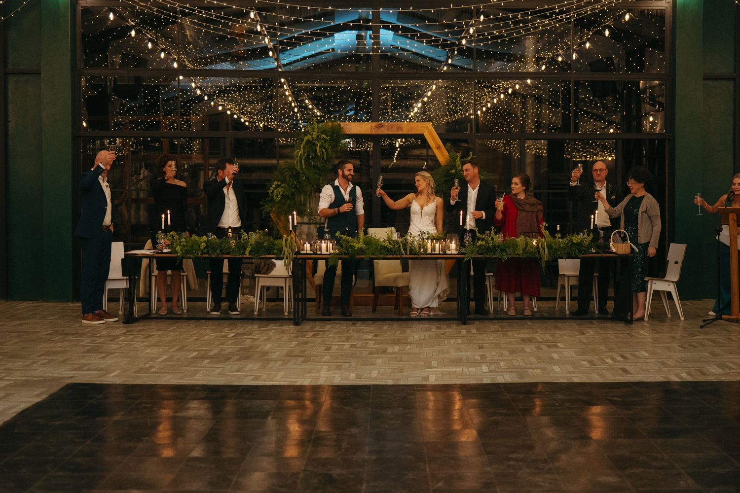 Wedding reception with bridal party at decorated head table, string lights, greenery, and hexagon backdrop in elegant venue.