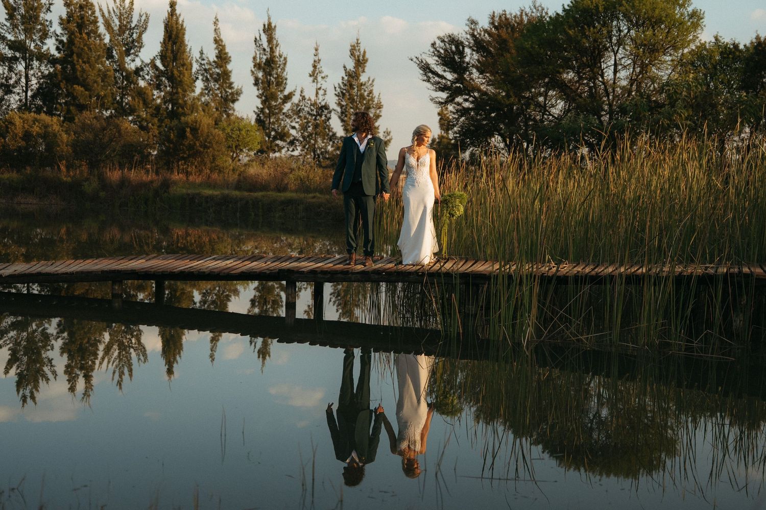 Bride and groom walking on a wooden dock reflected in calm water, surrounded by lush greenery at golden hour.
