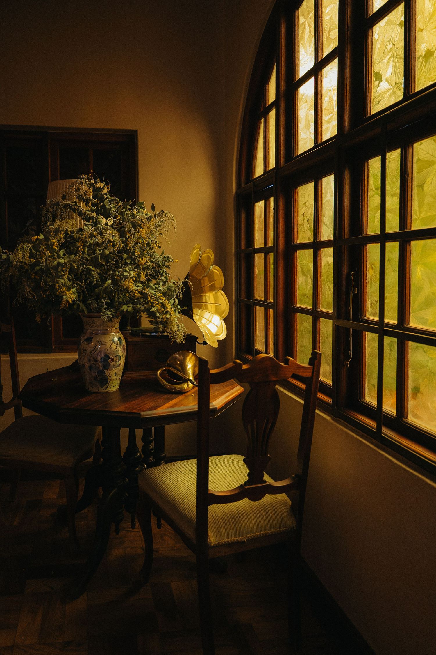 Cozy dimly lit corner with antique wooden table, chairs, floral vase, and glowing lamp by arched window.