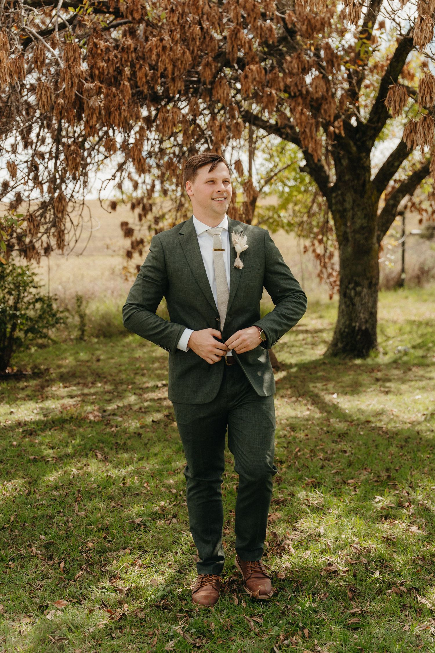 Groom in green suit with boutonniere stands outdoors under autumn tree on wedding day.