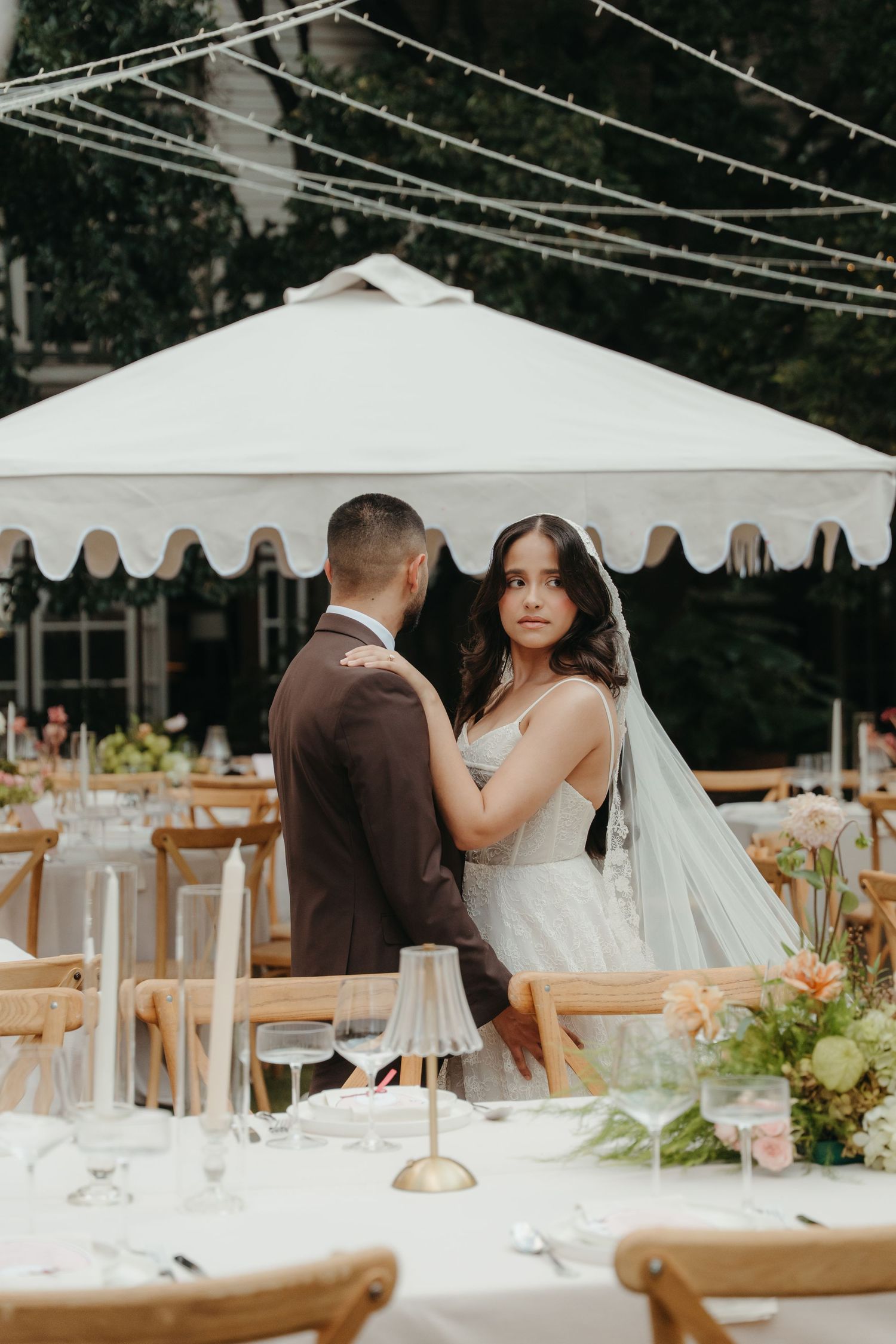 Bride and groom share a tender moment at an elegant outdoor wedding reception with string lights and white tent.
