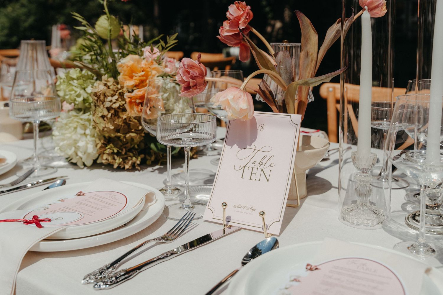 Elegant wedding table setting with floral centerpiece, crystal glasses, silverware, and a Table Ten sign at an outdoor reception.