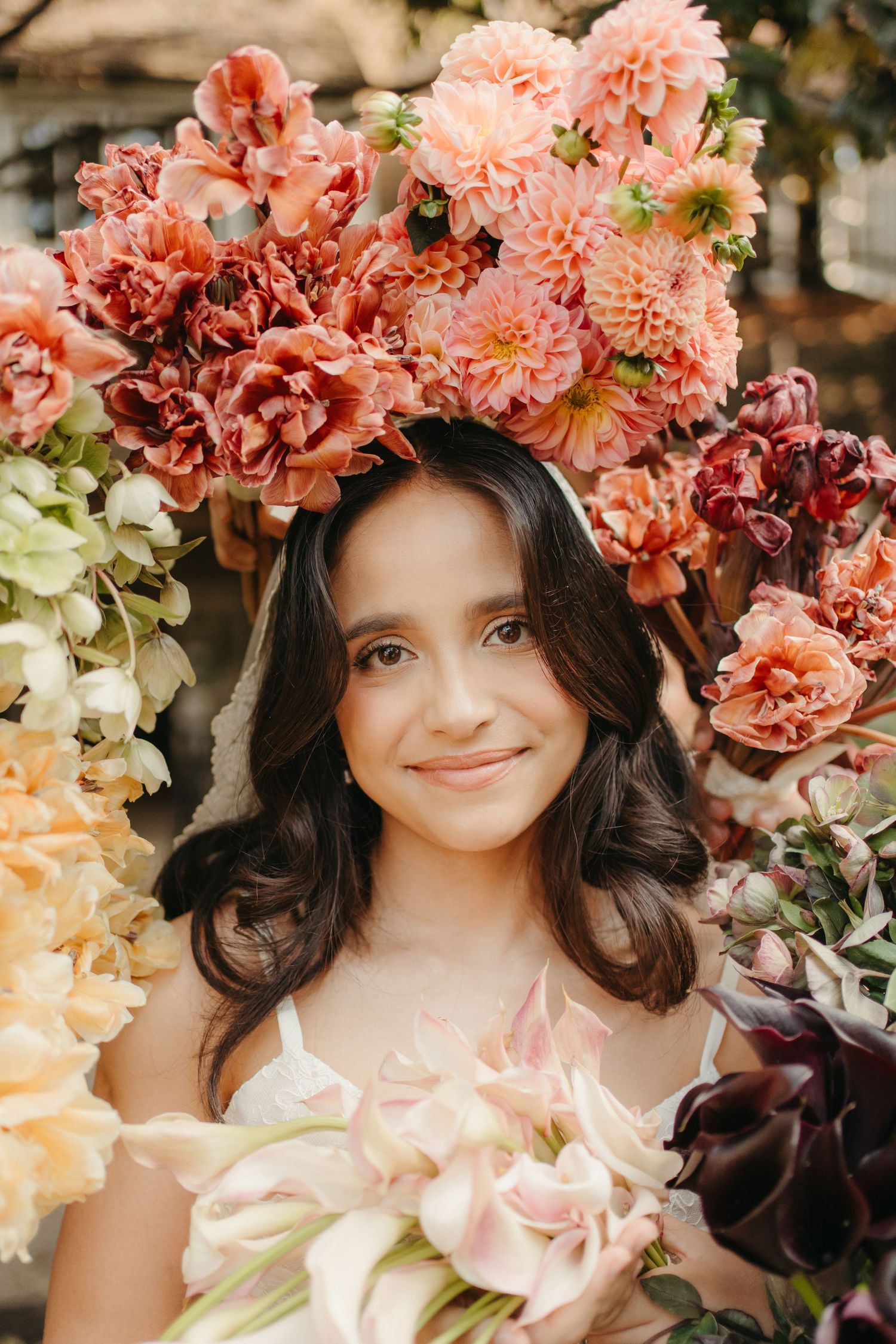 Young woman smiling surrounded by lush pink and coral floral arrangements including dahlias and carnations.