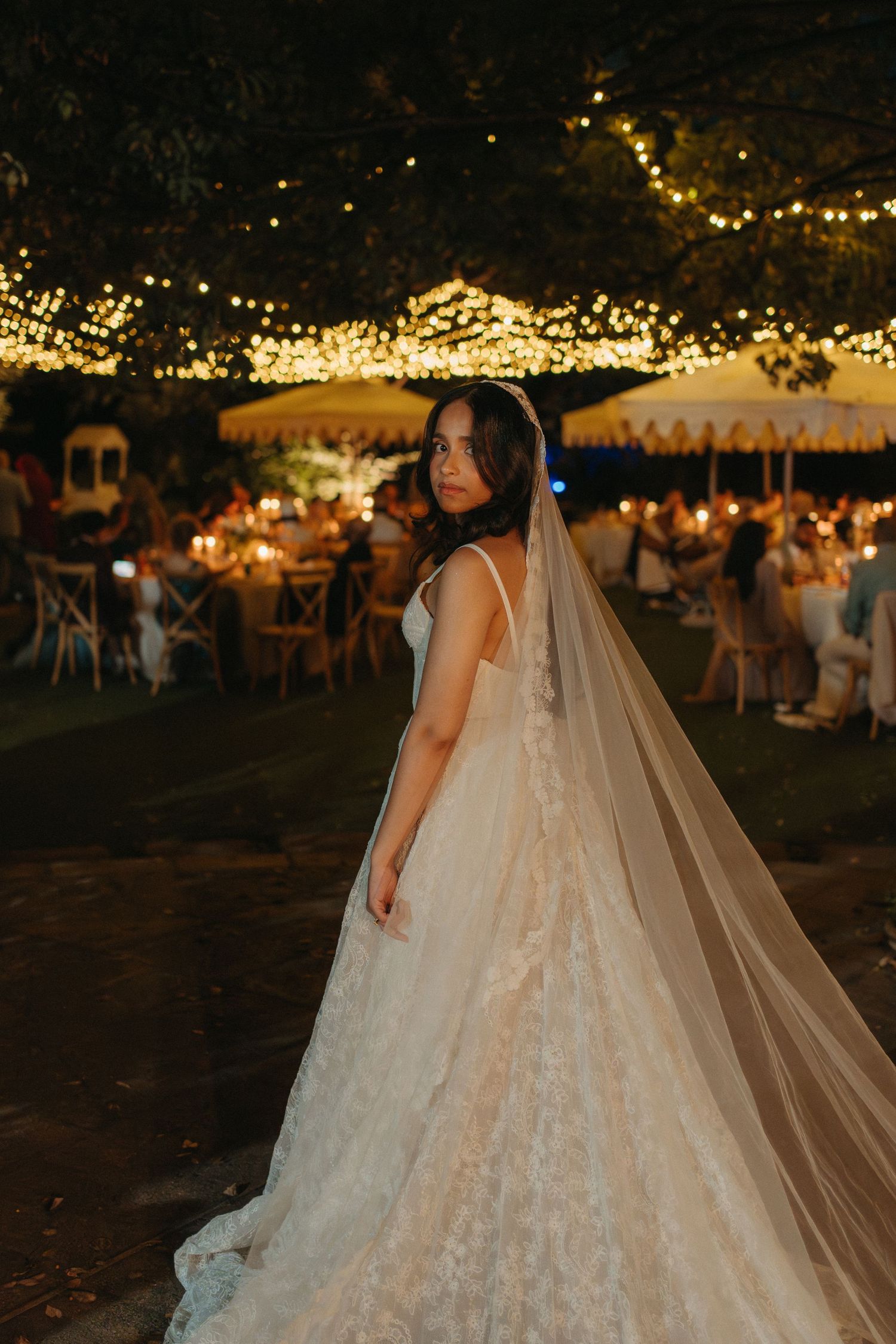 Bride in lace ballgown with flowing veil poses at outdoor evening wedding reception with string lights.