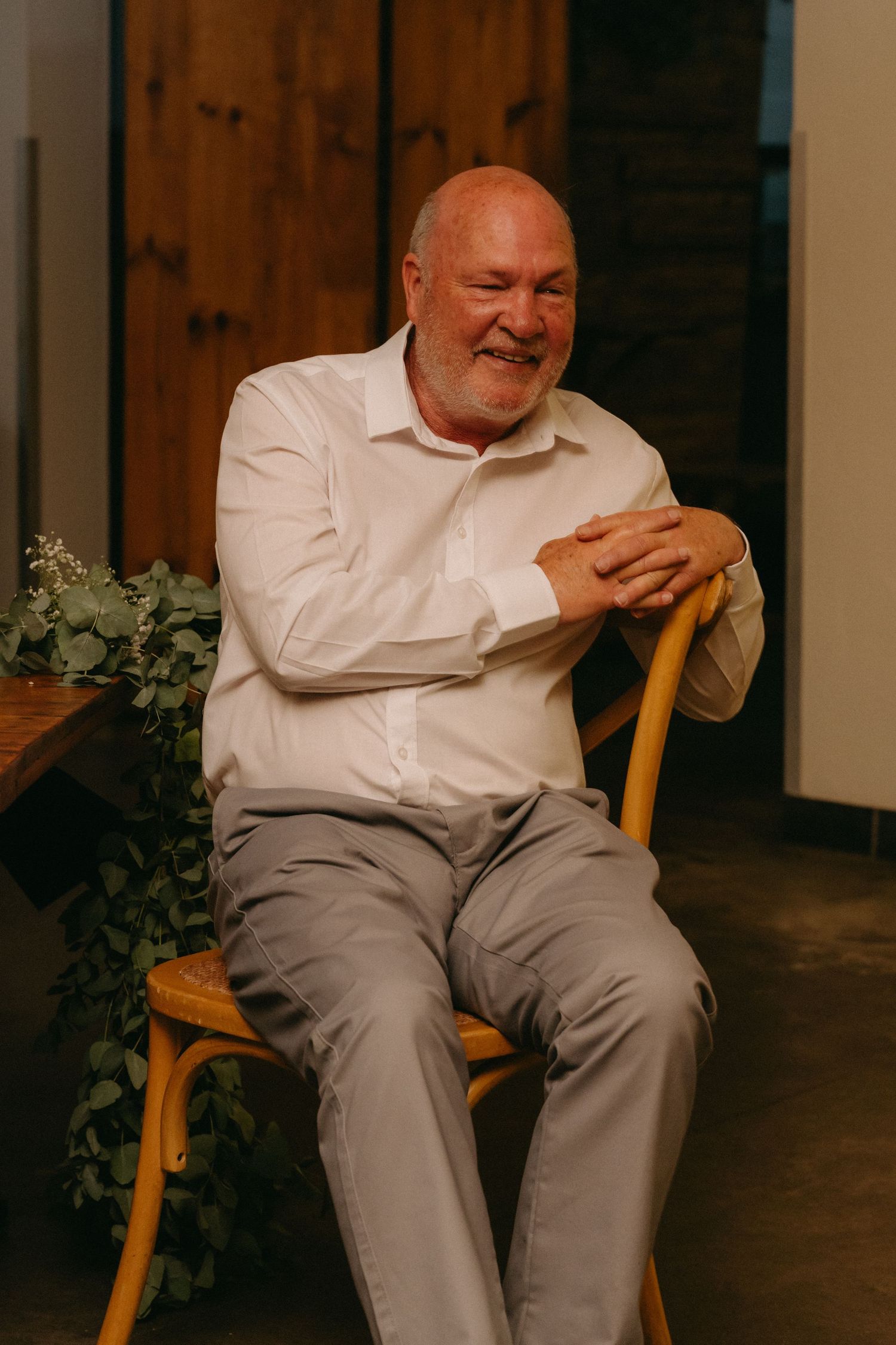 An older gentleman in white shirt and gray pants smiles while seated on a wooden chair.