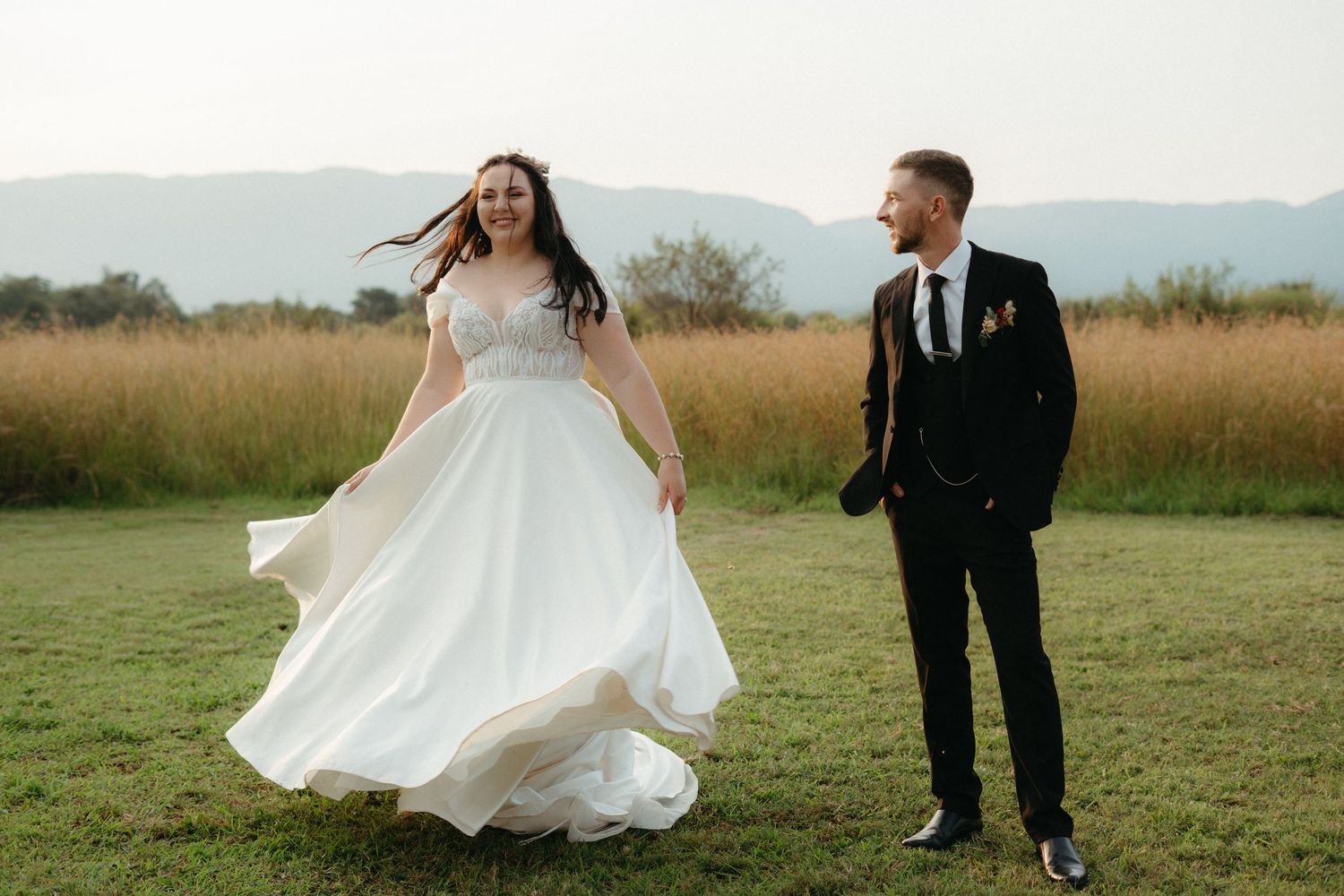 A bride's dress flows in the wind as she dances in a field while her groom watches.