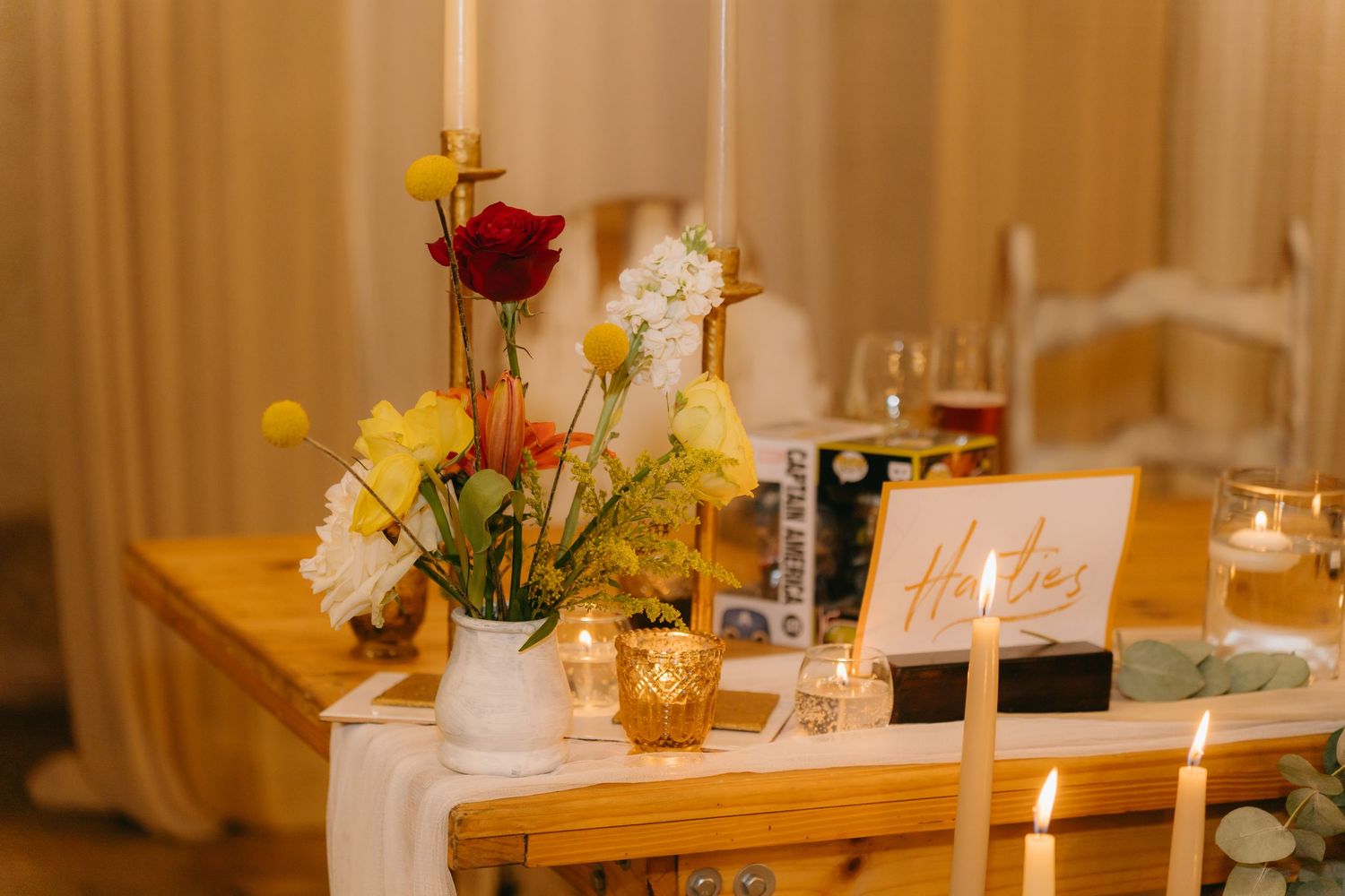 Wedding reception table with red and yellow flowers in vase, candles and decorative elements.