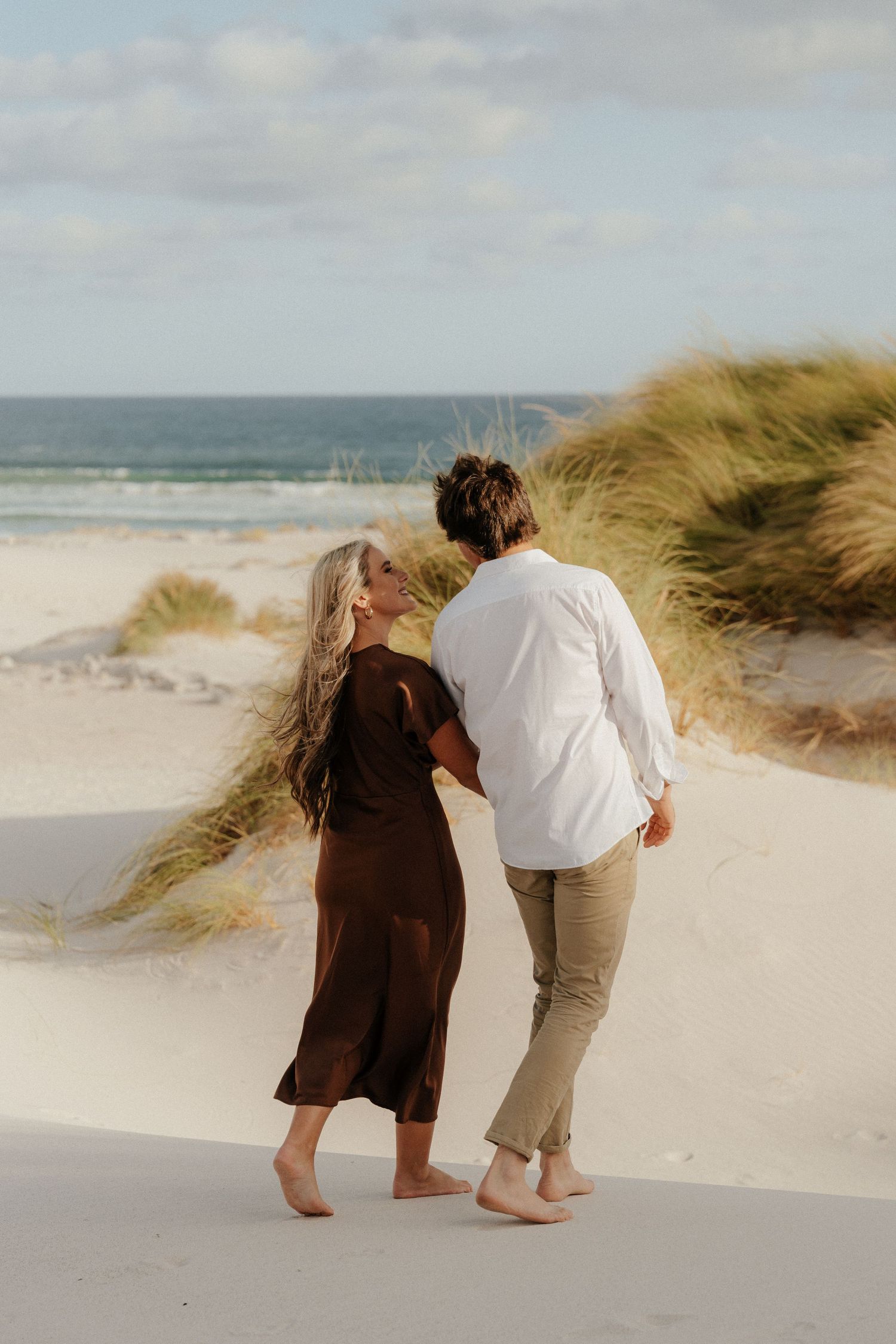 Couple walking barefoot on white sand dunes near the ocean, woman in brown dress and man in white shirt.