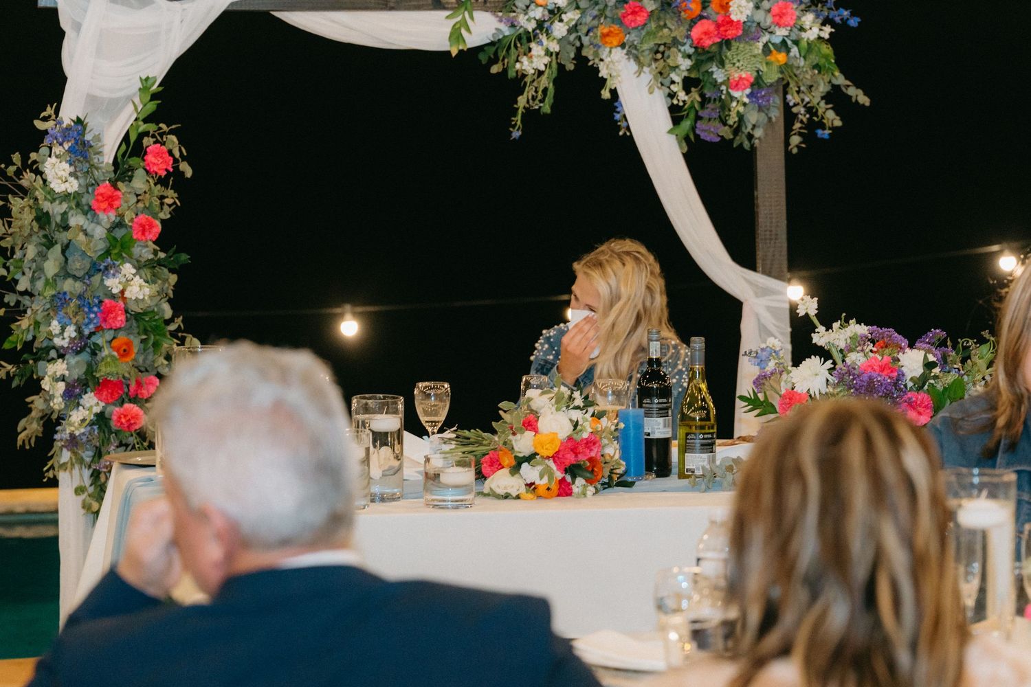 An outdoor wedding reception setup with floral arch decorations and string lights at night.