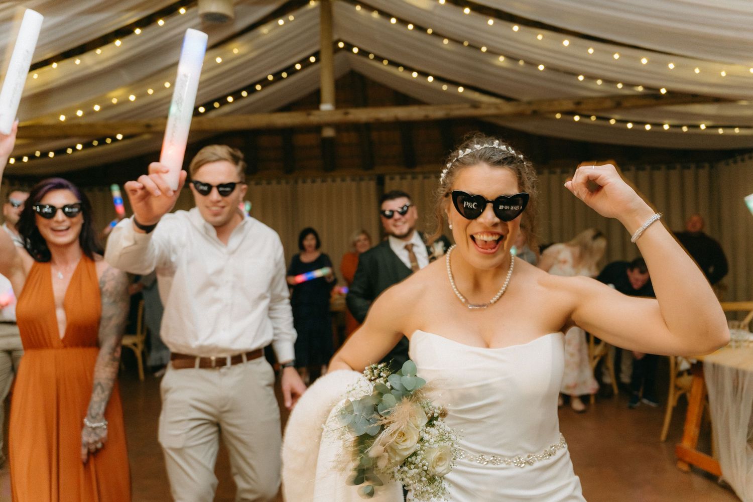Wedding party dancing and celebrating on the dance floor under string lights at an indoor reception venue.