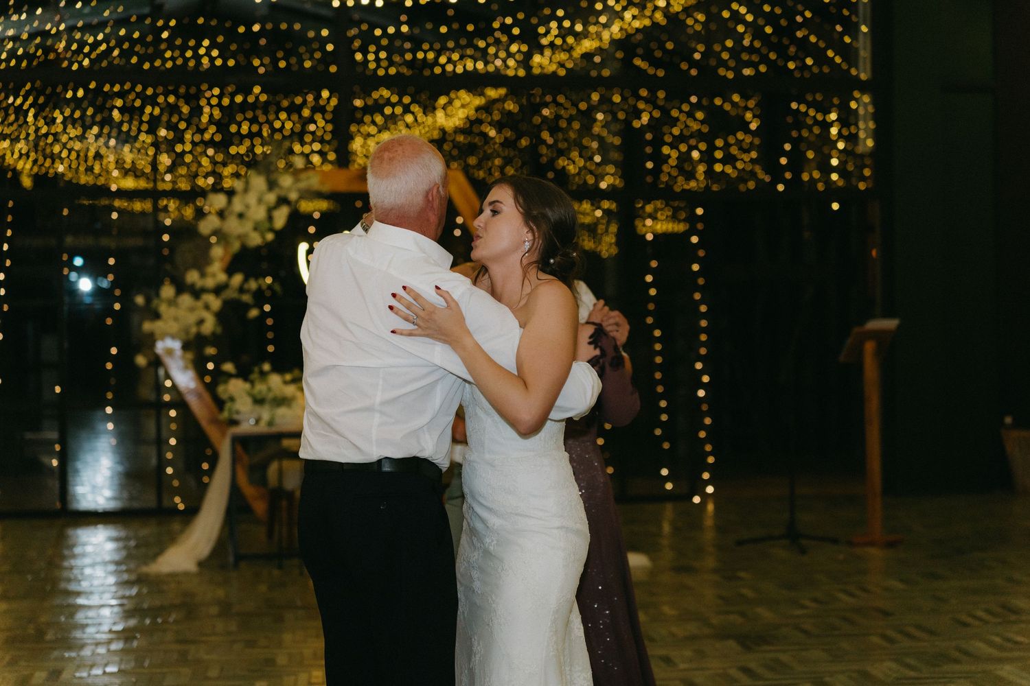 Couple shares intimate dance moment under warm string lights at wedding reception.