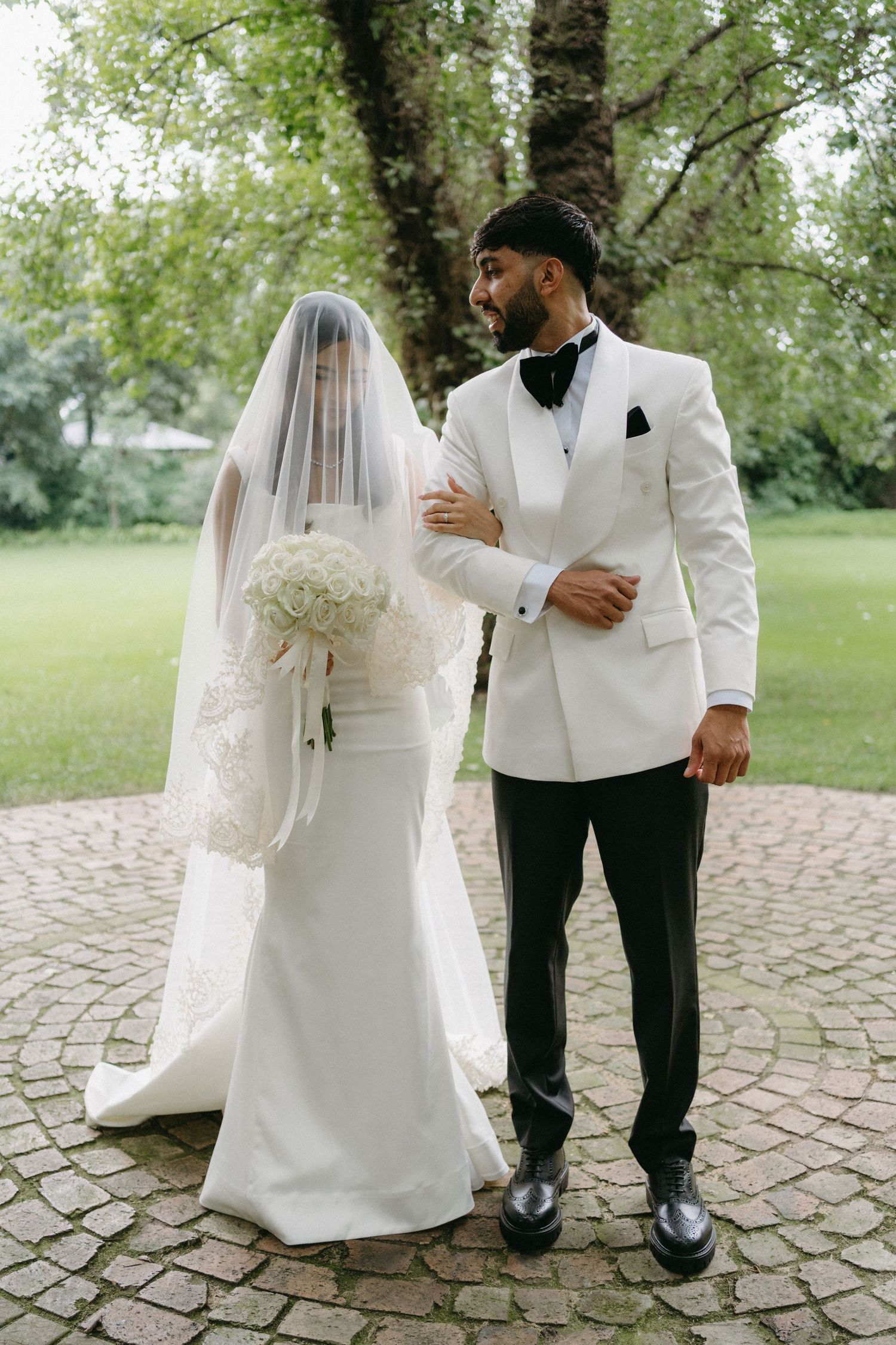 A wedding couple stands on a circular stone patio surrounded by lush greenery, with the groom in a white tuxedo jacket.