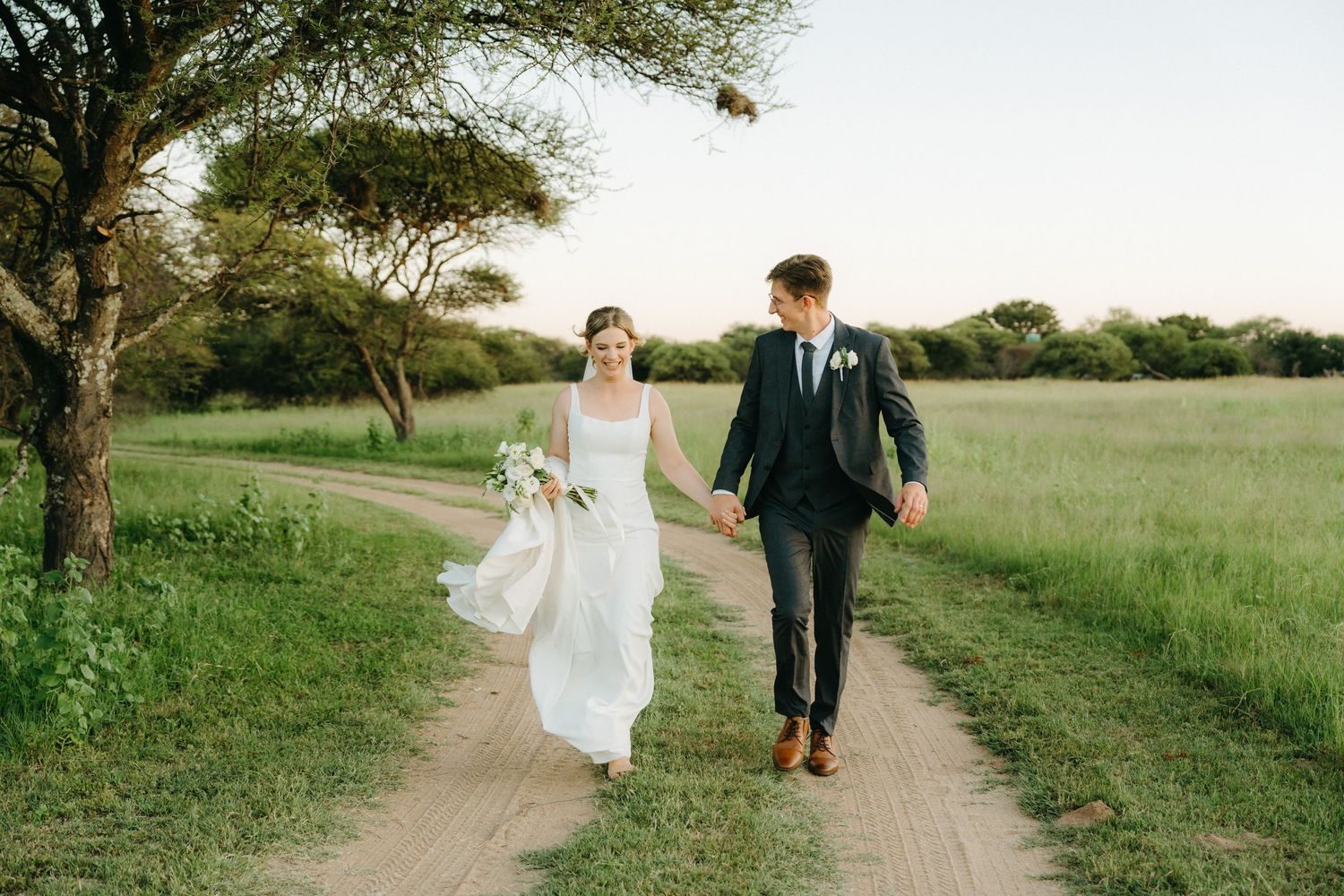 A couple walks hand in hand on a dirt path through a green field on their wedding day.