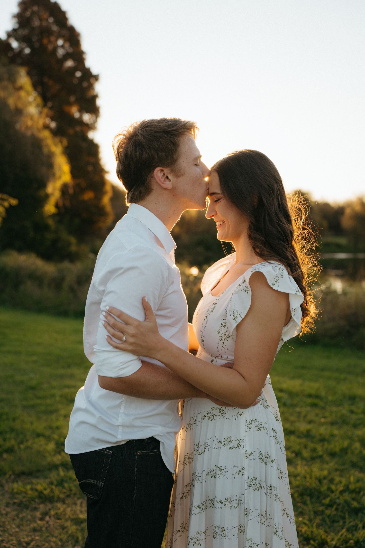 Romantic couple embraces during golden hour sunset in a grassy field with trees in the background.