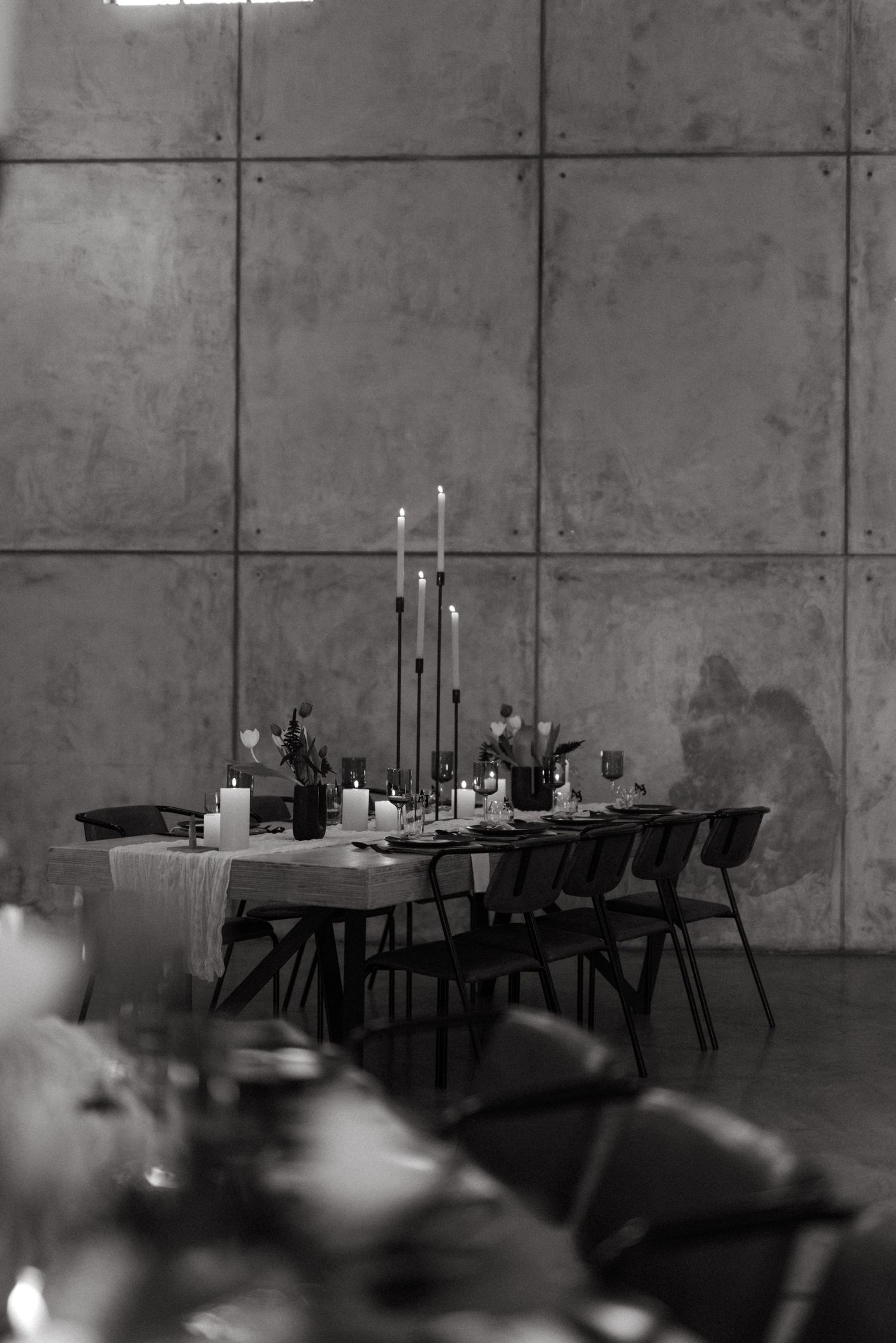 Elegant table setting with tall candles and black chairs against concrete wall in moody black and white photography.
