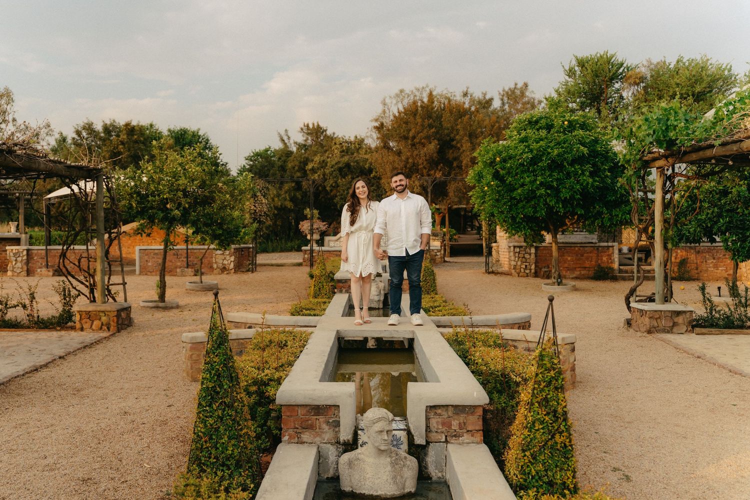 Couple walking through a rustic greenhouse with wooden tables and natural light streaming through glass panels.