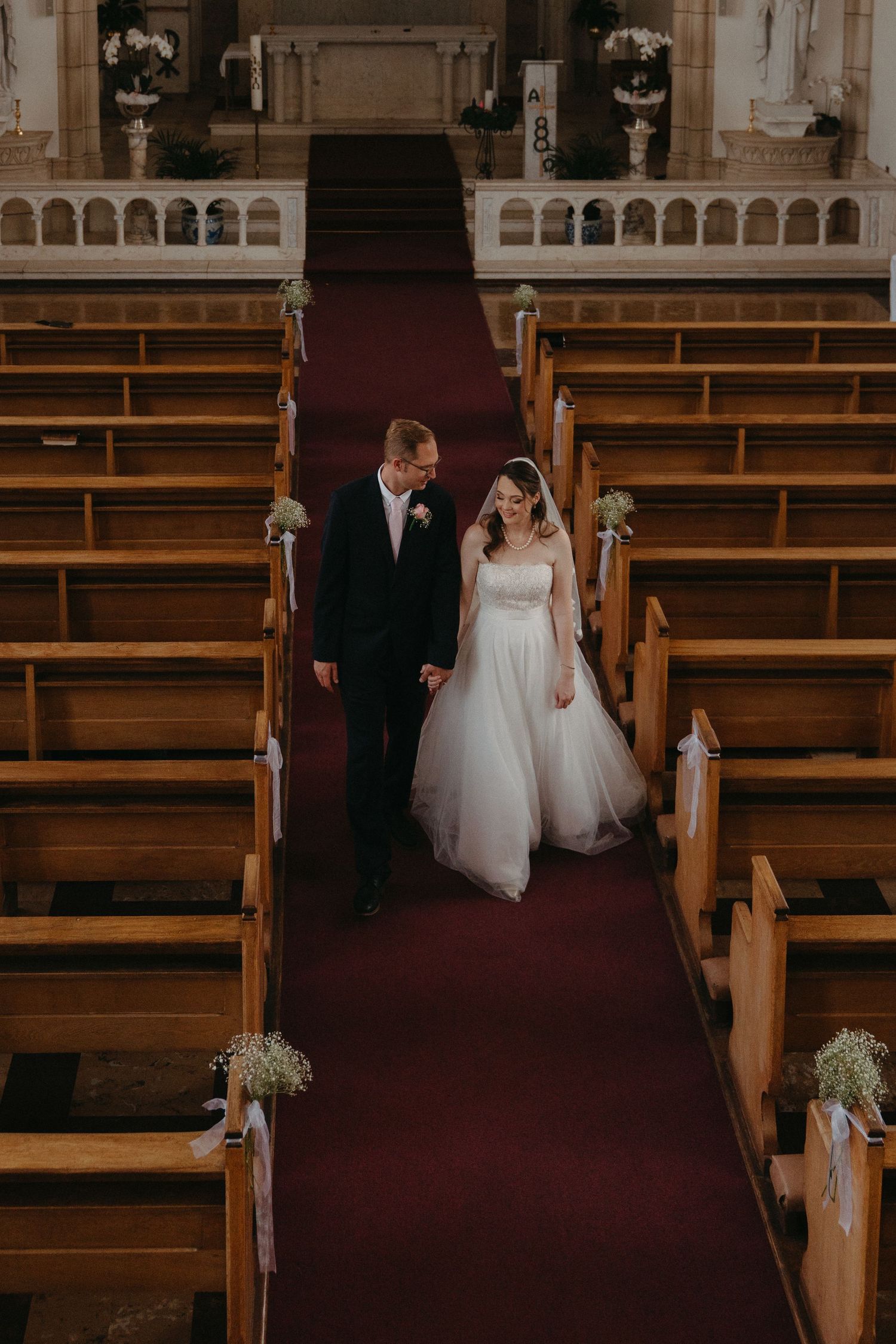 A bride and groom walk down the aisle of a traditional church with wooden pews decorated with white floral arrangements.