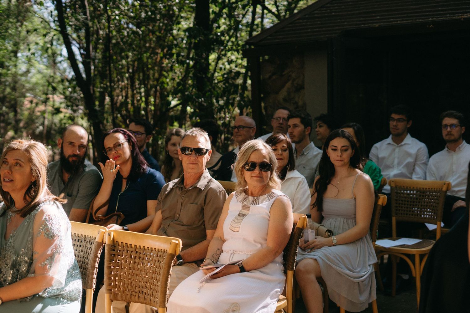 Wedding guests sitting on rattan chairs enjoy an outdoor ceremony in dappled sunlight.