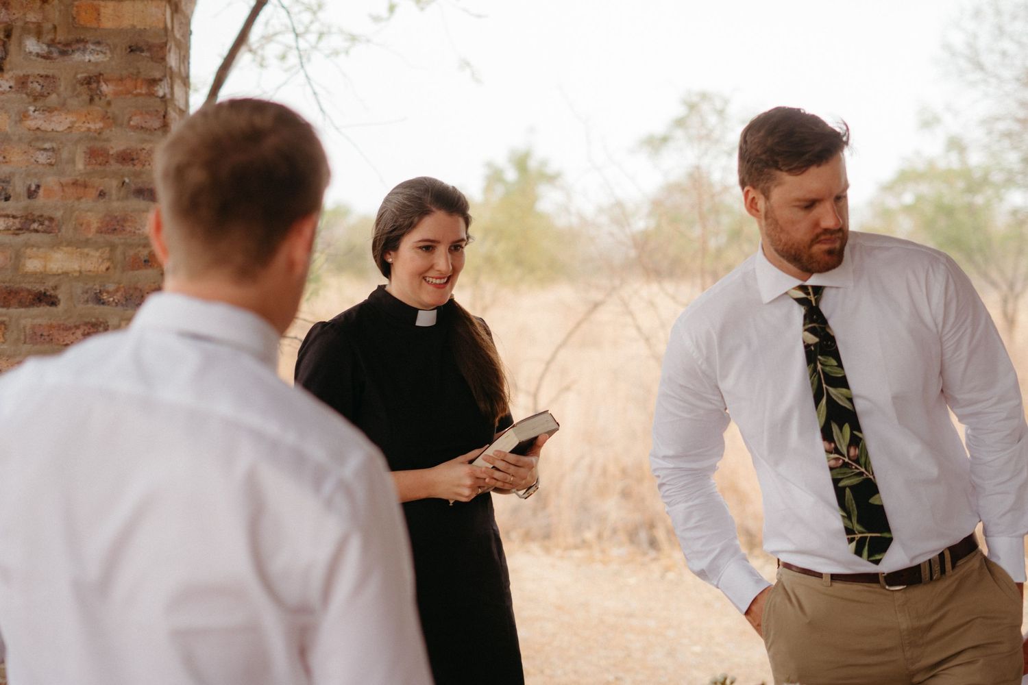 A casual outdoor wedding ceremony with attendees gathered under a rustic brick structure.