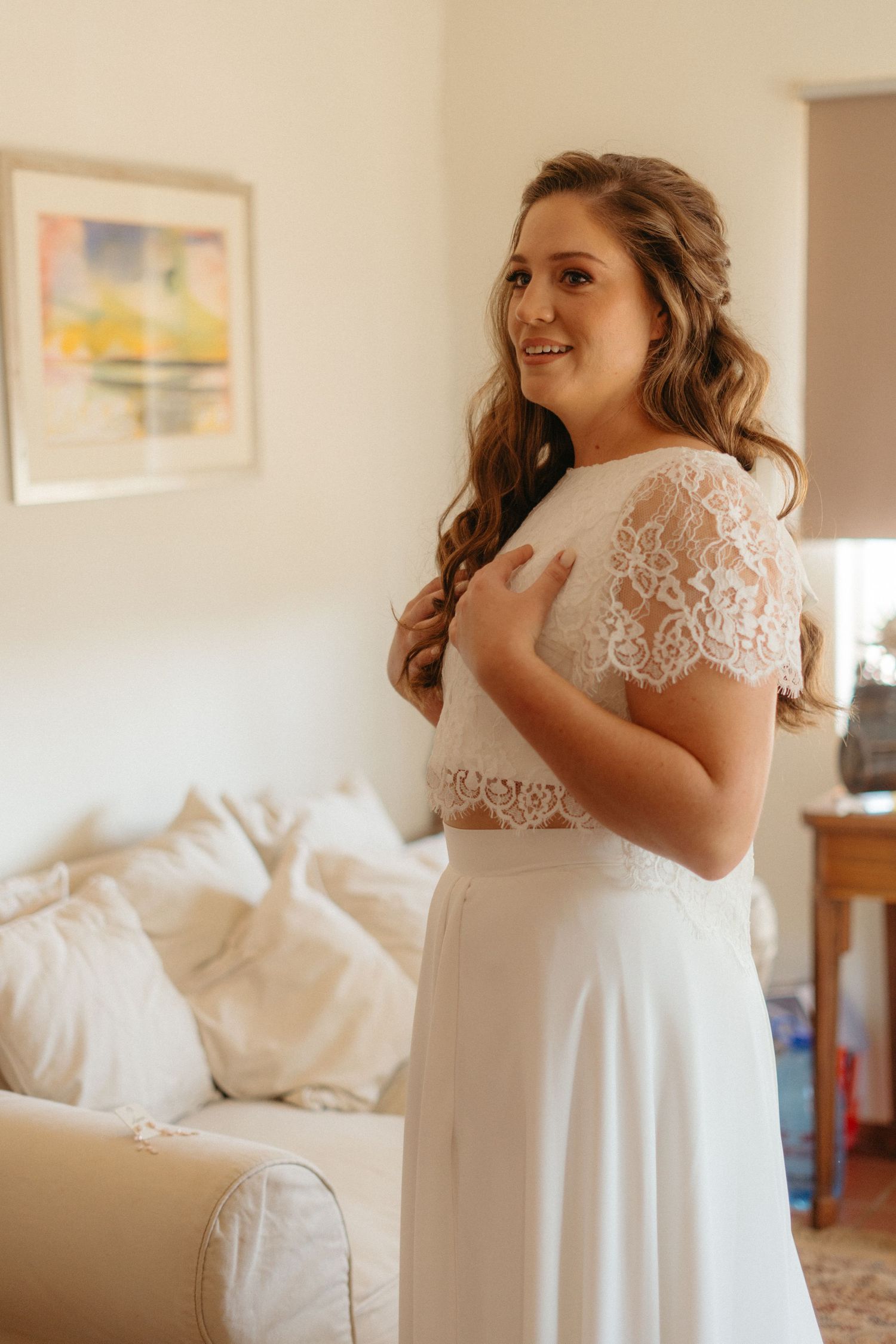 A bride in a two-piece lace wedding dress smiling in a sunlit room.