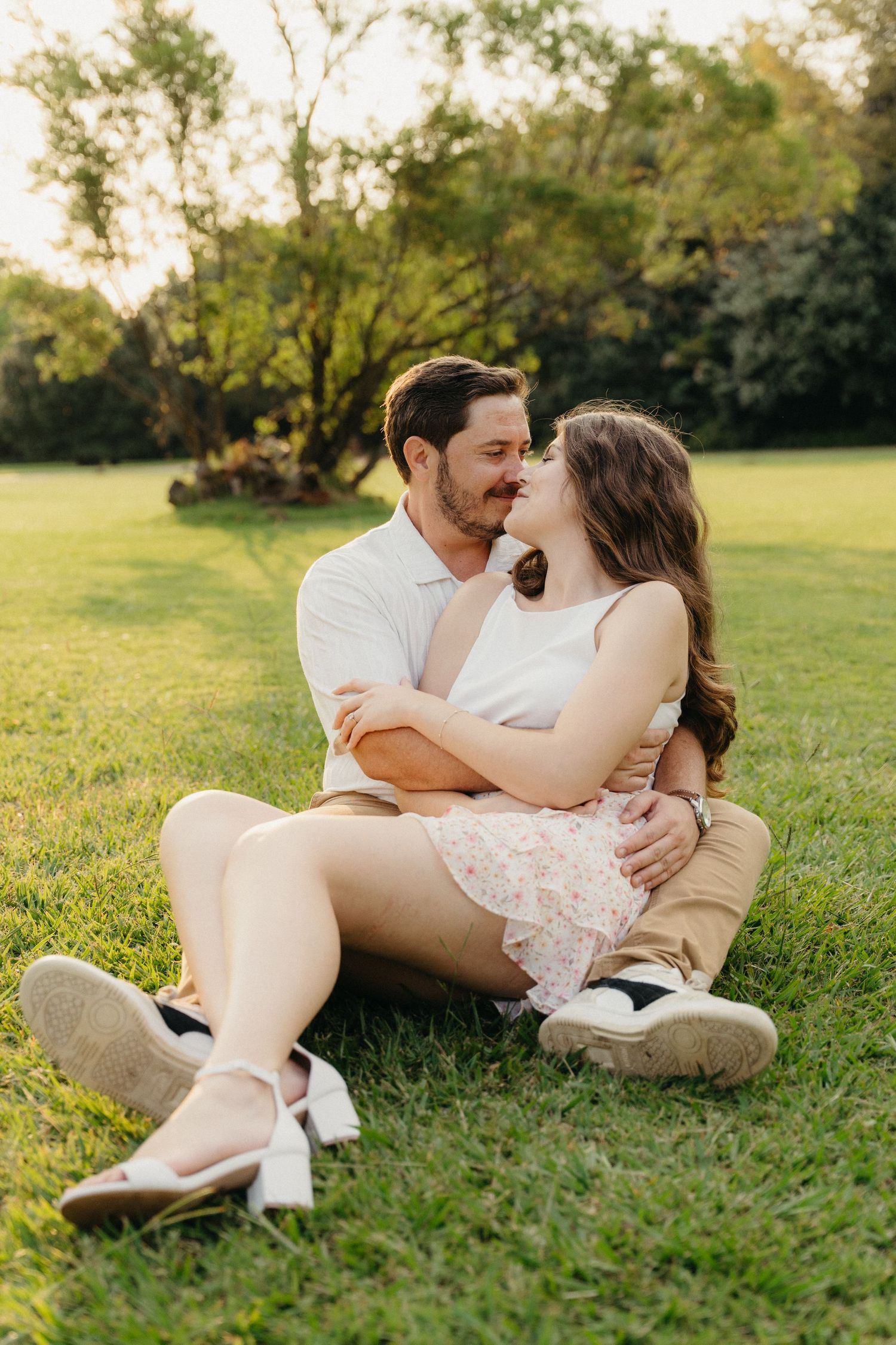 A couple sits on green grass in a park, embracing and about to kiss in warm golden hour sunlight.