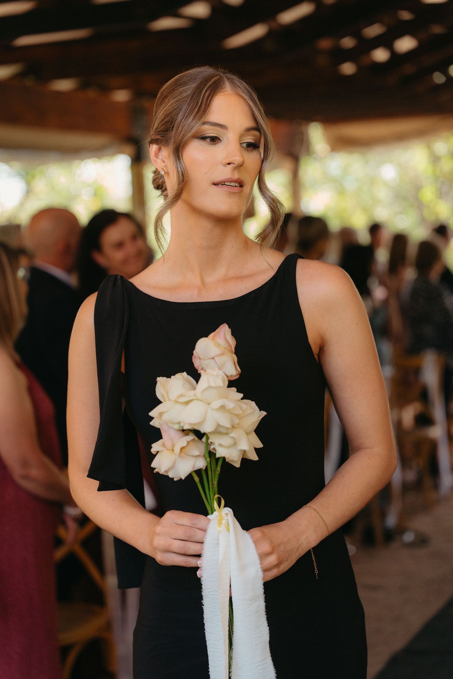 A bridesmaid in a black dress holds white flowers with ribbons at an indoor wedding venue.
