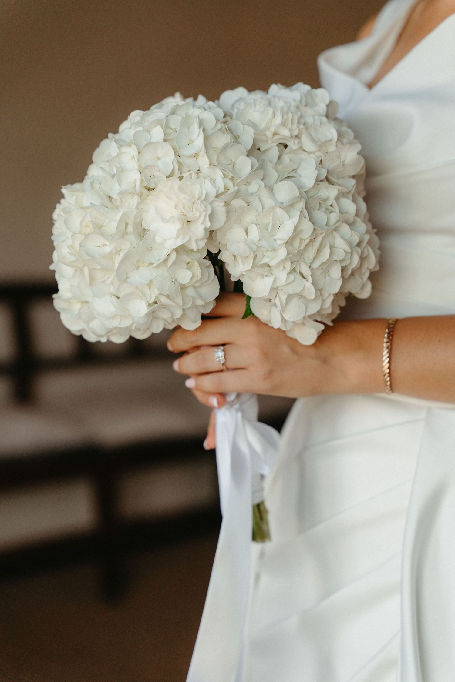 A bride in a white dress holds an elegant bouquet of white hydrangea flowers against a soft background.