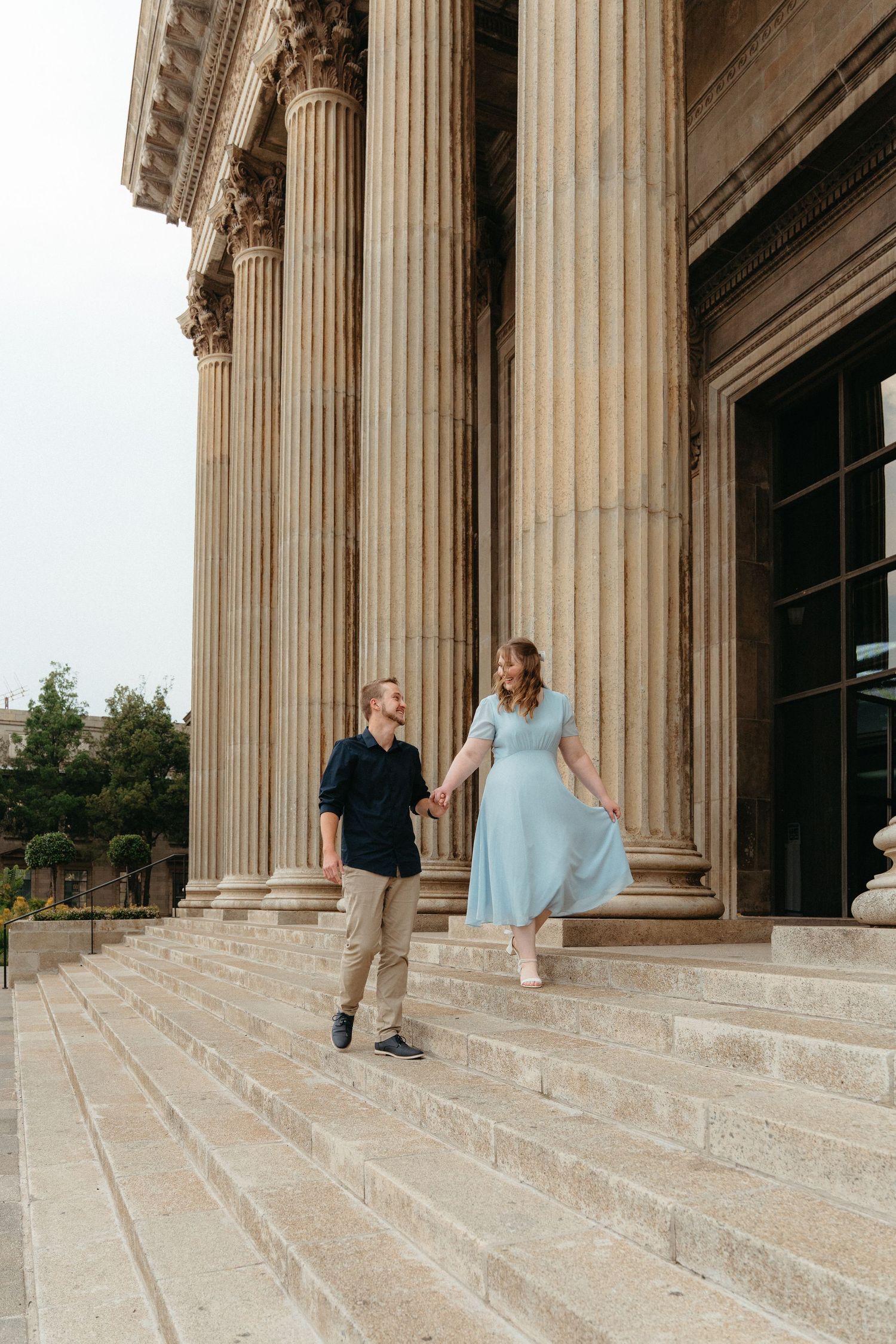 A couple in formal attire poses together on grand stone steps flanked by classical columns at a historic building.