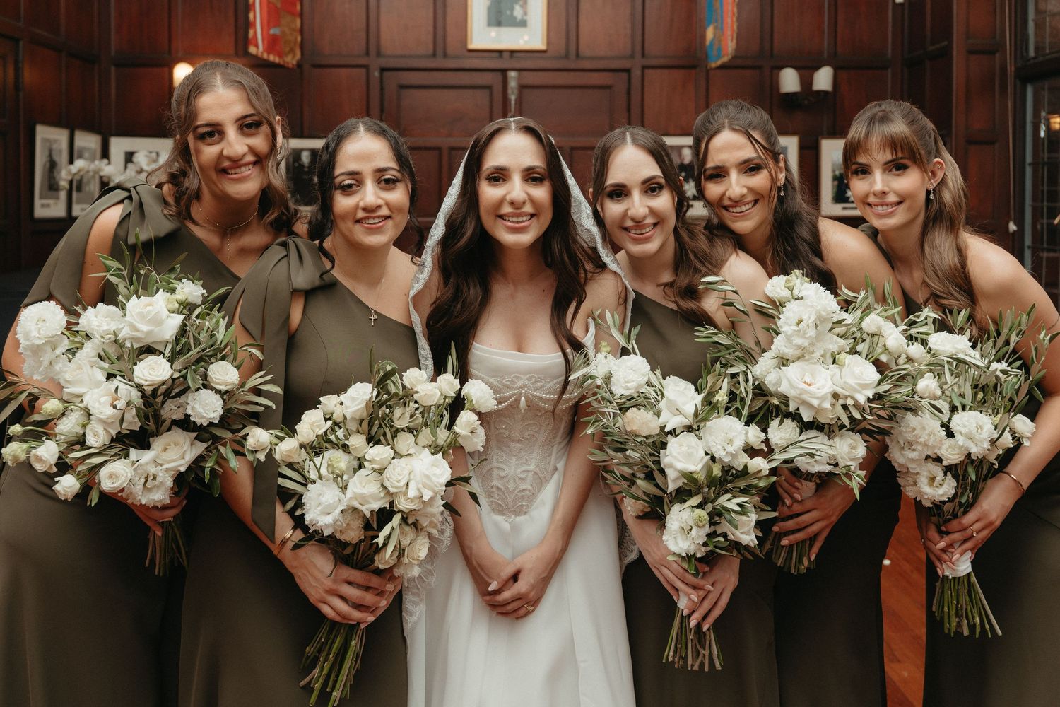 Bride in white gown with five bridesmaids in olive green dresses holding white floral bouquets posing together indoors.