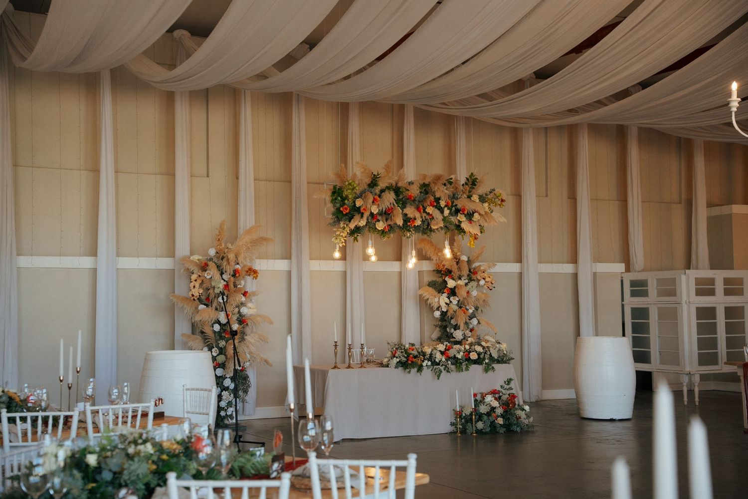Elegant wedding reception venue with draped ceiling fabric, a floral arch backdrop, a white head table, and candlelit tables with greenery.