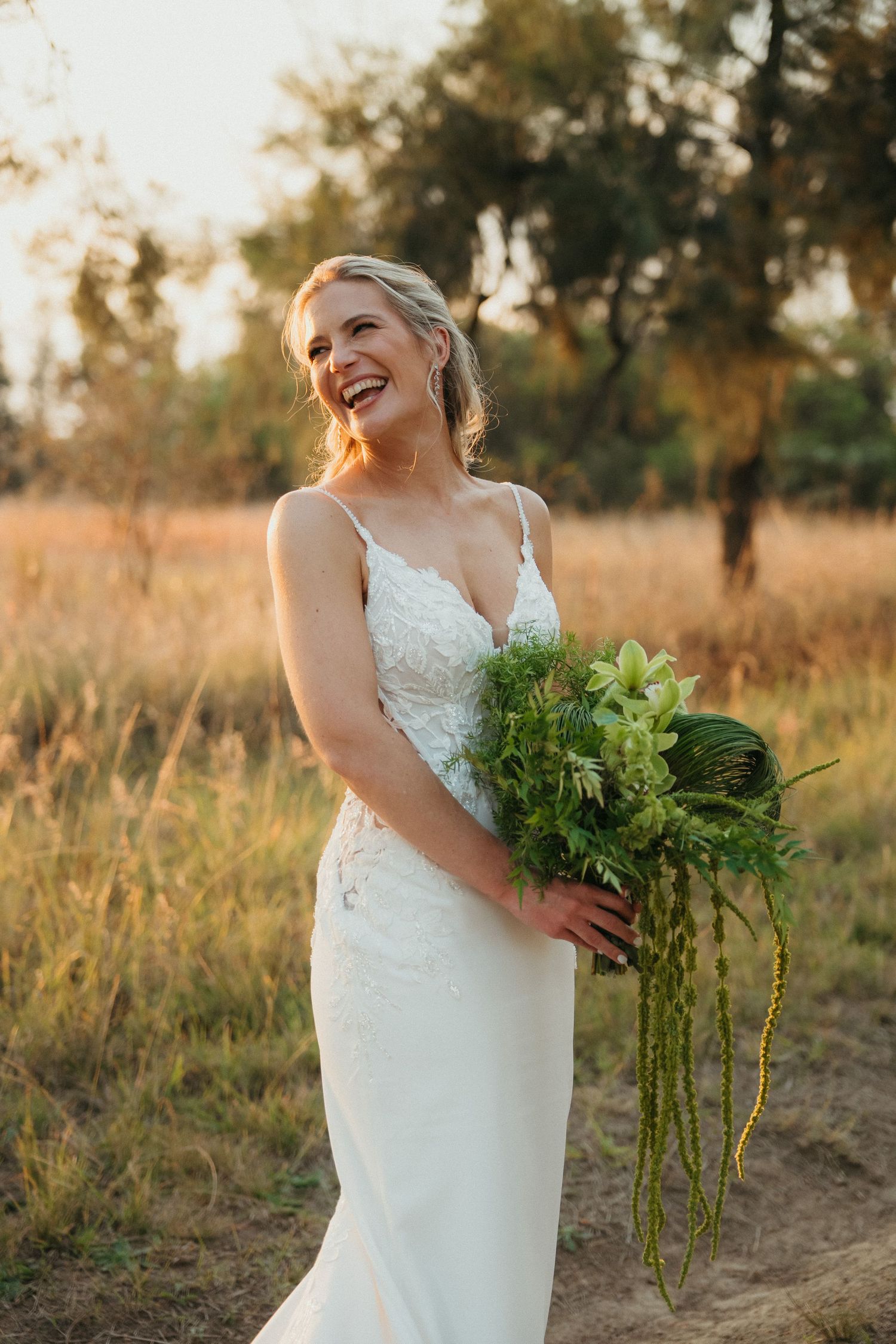 Laughing bride in white lace gown holds green bouquet of ferns outdoors at golden hour.