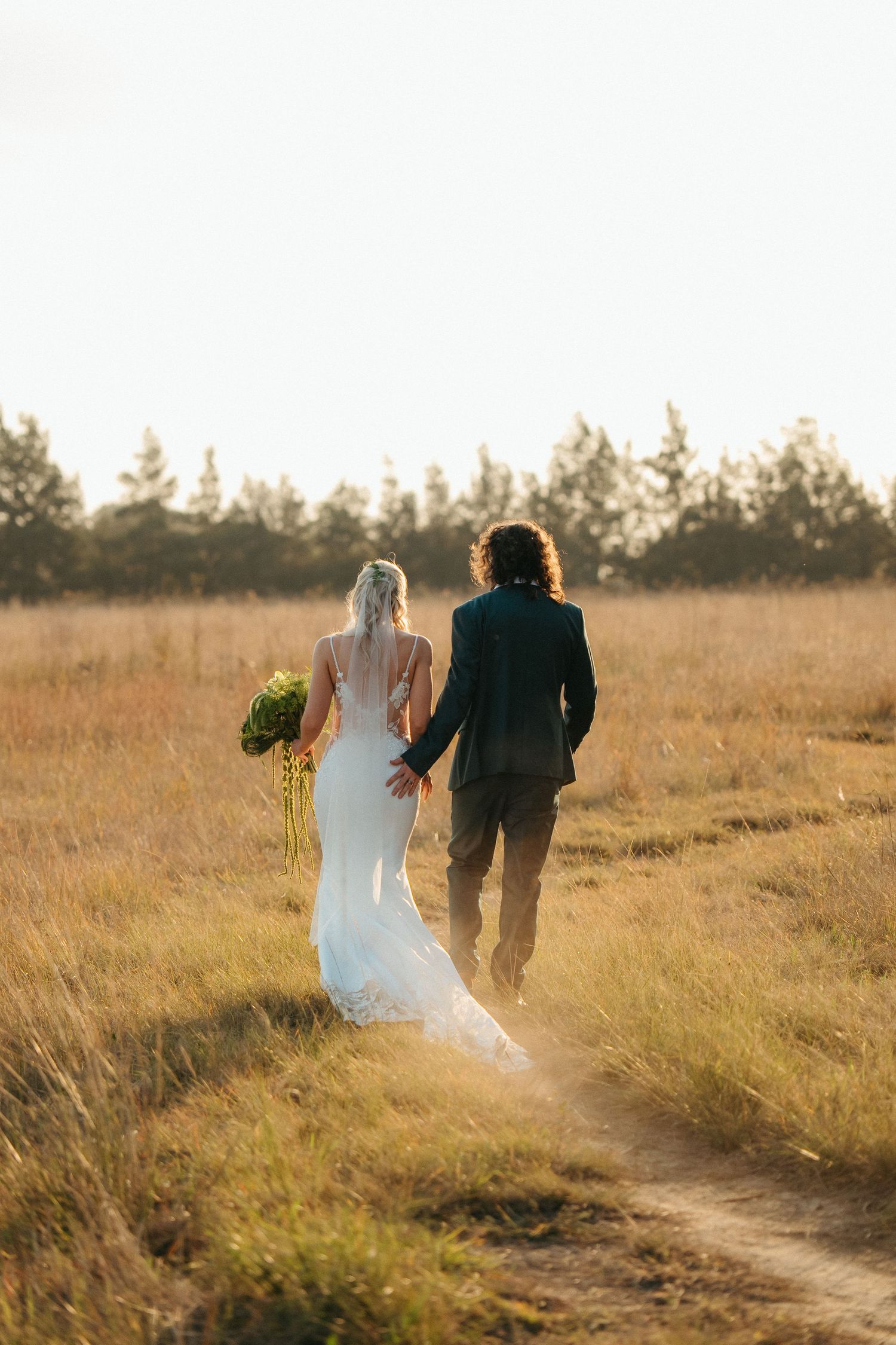 Bride and groom walking hand in hand through golden field at sunset, bride holding bouquet in white gown.
