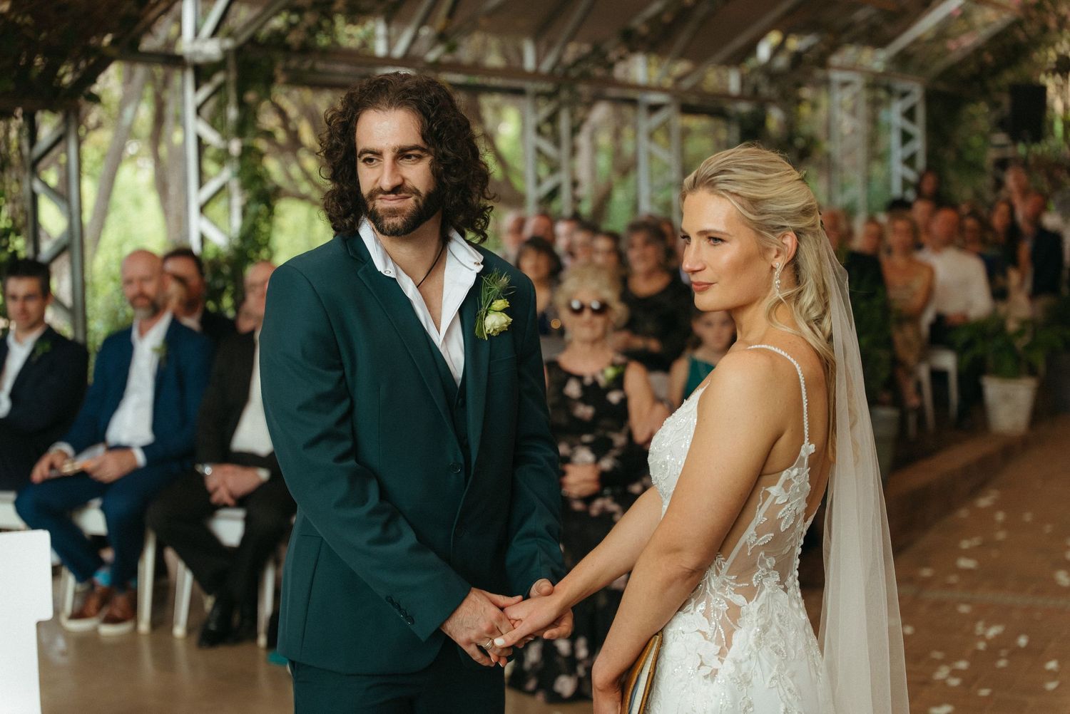 Bride and groom holding hands at outdoor wedding ceremony, groom in green suit, bride in lace gown with veil.