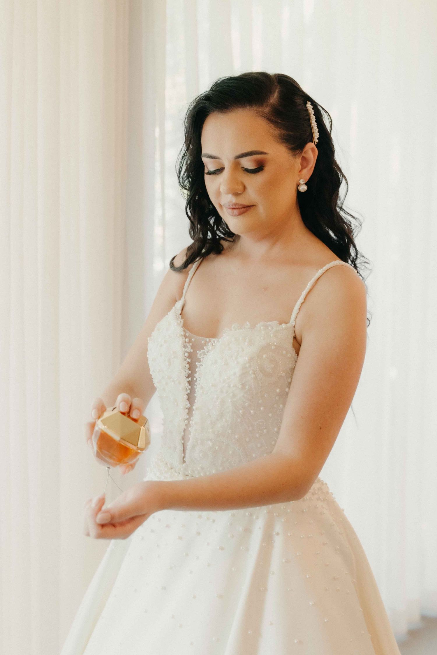A bride in a beaded white gown applies perfume while getting ready for her wedding day.