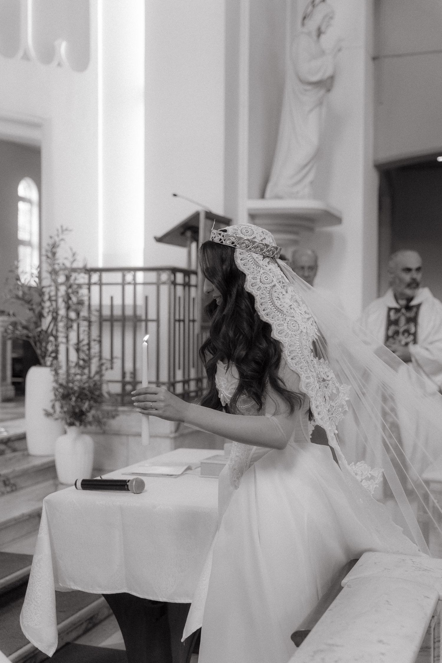 Black and white photo of a bride in lace veil kneeling at church altar during wedding ceremony with priest in background.