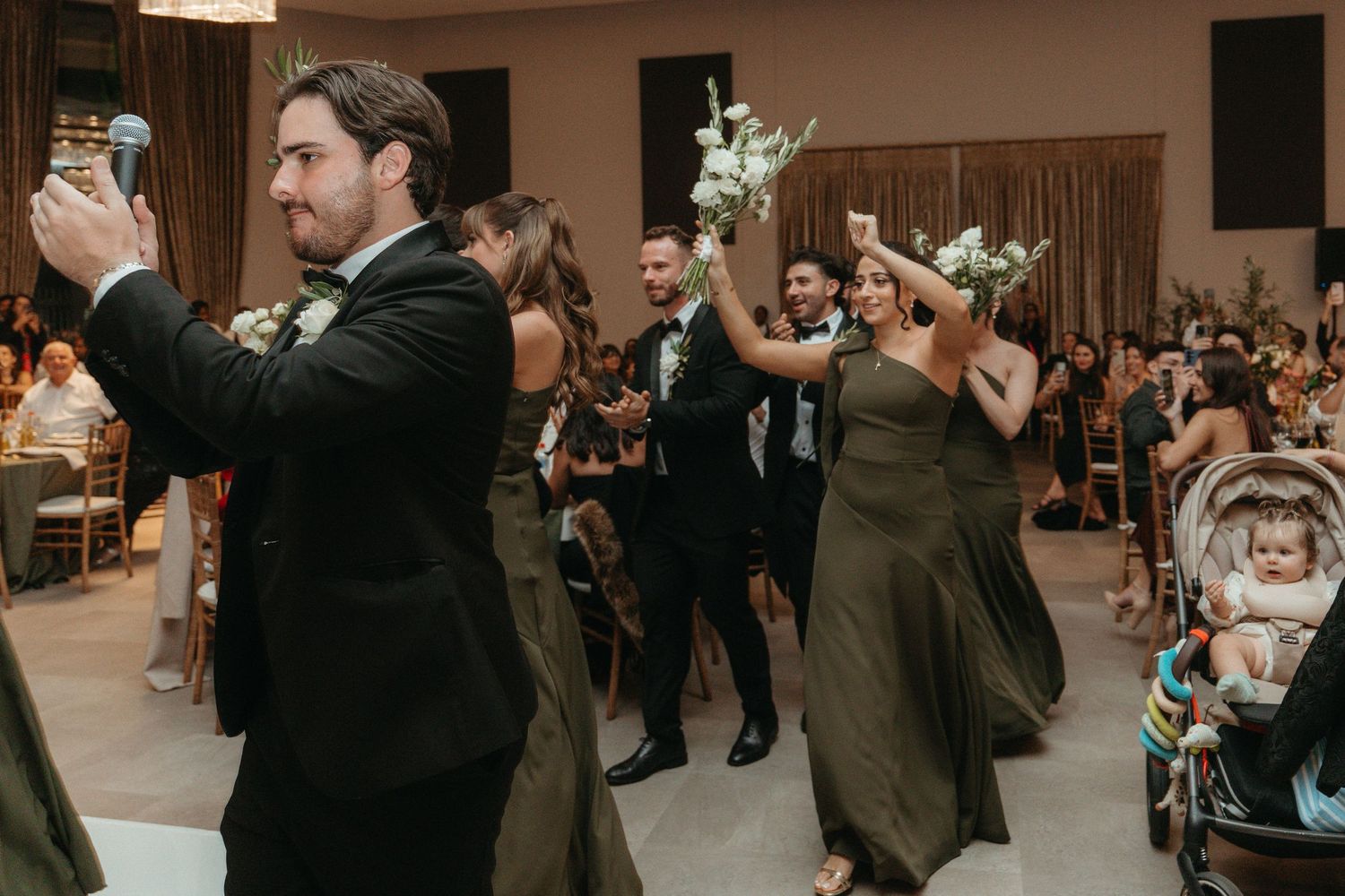 Wedding party processional with groomsmen and bridesmaids in olive dresses raising bouquets while guests watch in decorated reception hall.