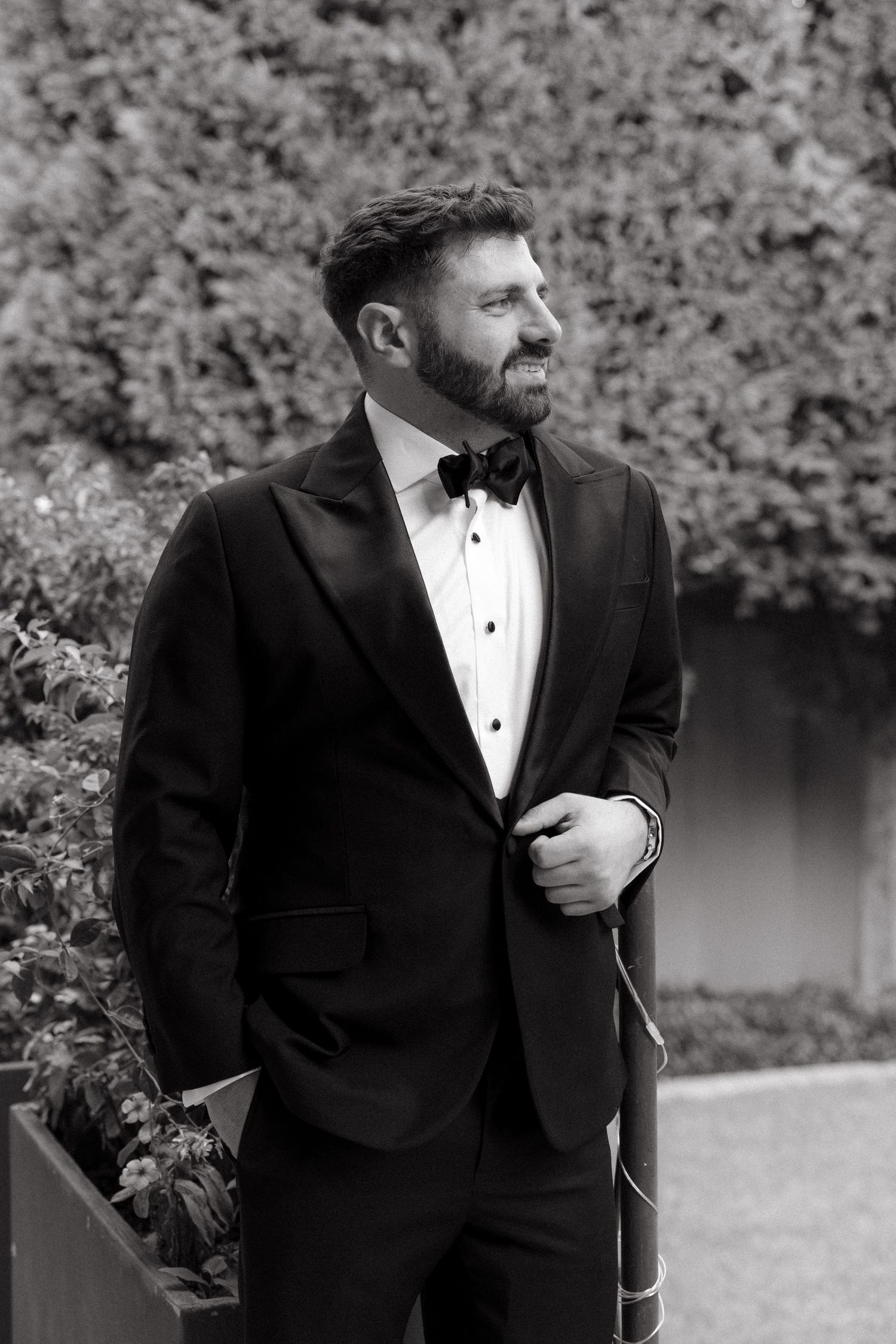 A bearded groom in a black tuxedo with bow tie smiles outdoors, posing confidently against lush green hedges in Johannesburg