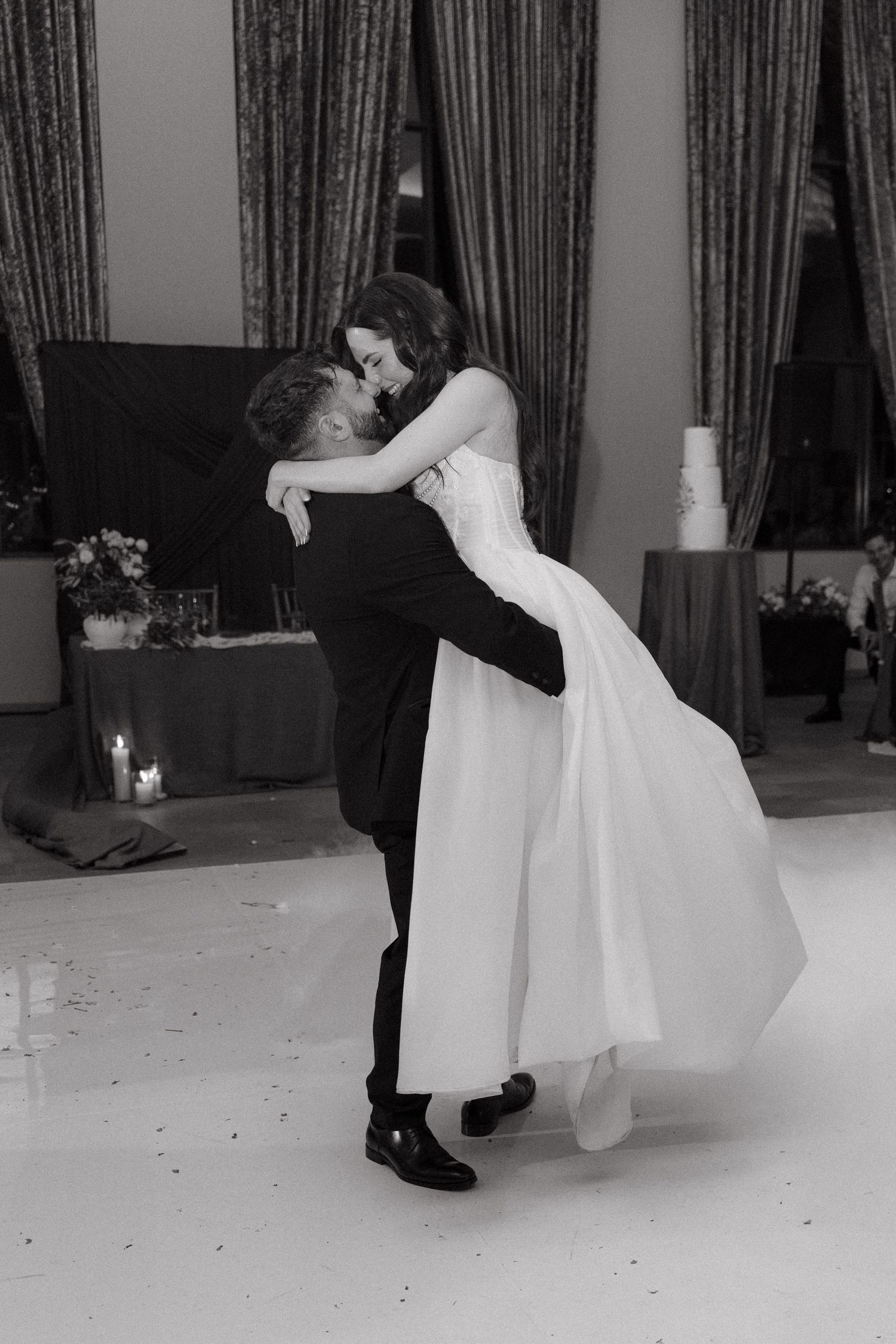 Black and white photo of groom lifting bride during romantic first dance at elegant wedding reception.